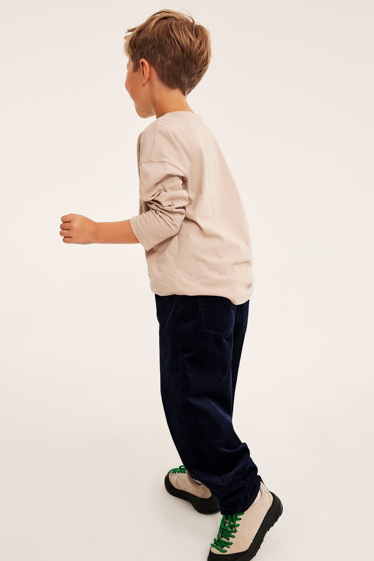 Sidovy av ett barn i mörkblå manchesterbyxor, en beige långärmad t-shirt och beiga sneakers.