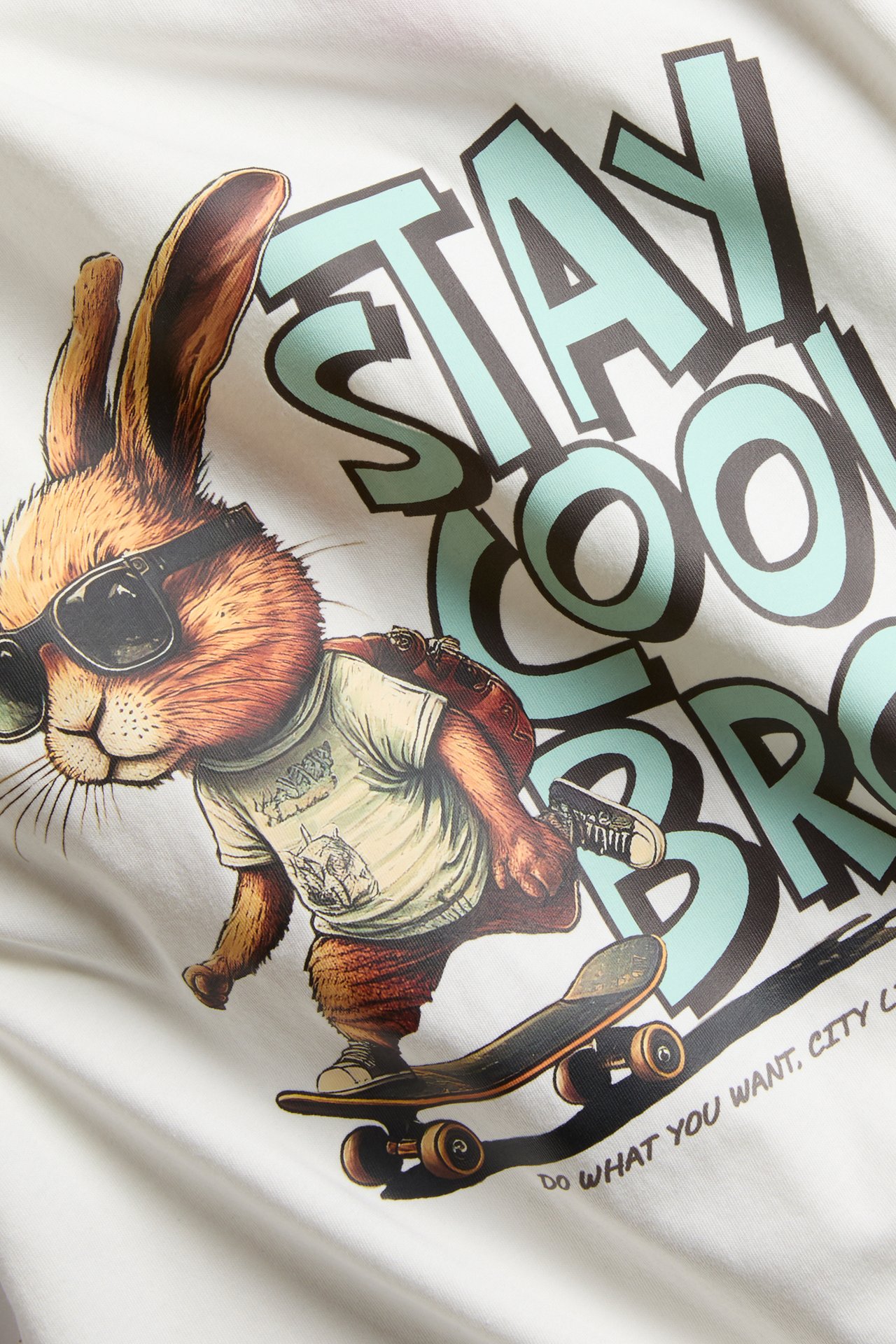 Lähikuva luonnonvalkoisesta puuvillaisesta t-paidasta, jossa on rullalautaileva kani ja teksti 'STAY COOL BRO'.