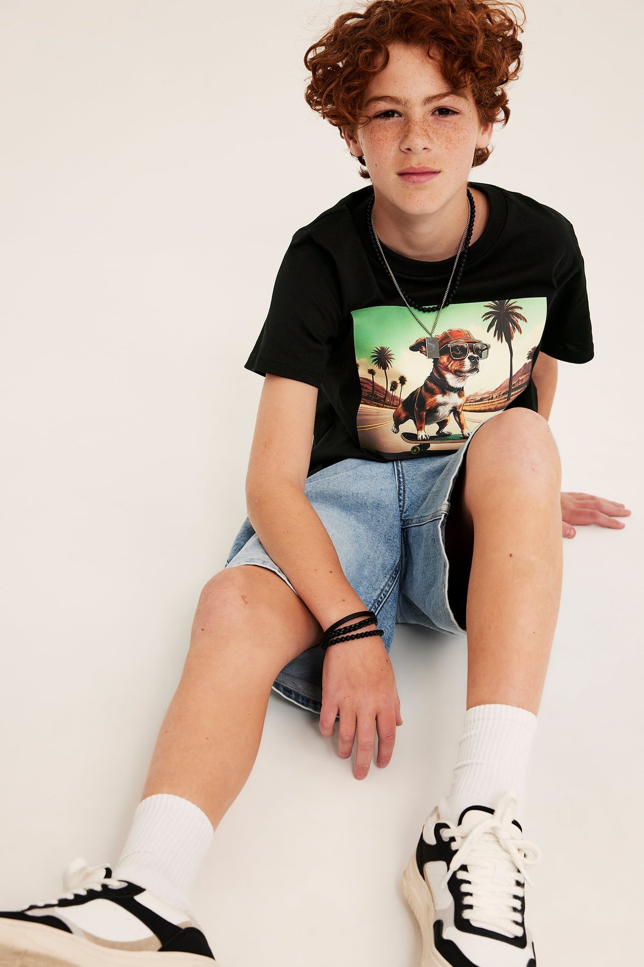 Frontvy på ett barn i svart t-shirt med hundtryck, denimshorts, halsband, armband och sneakers.