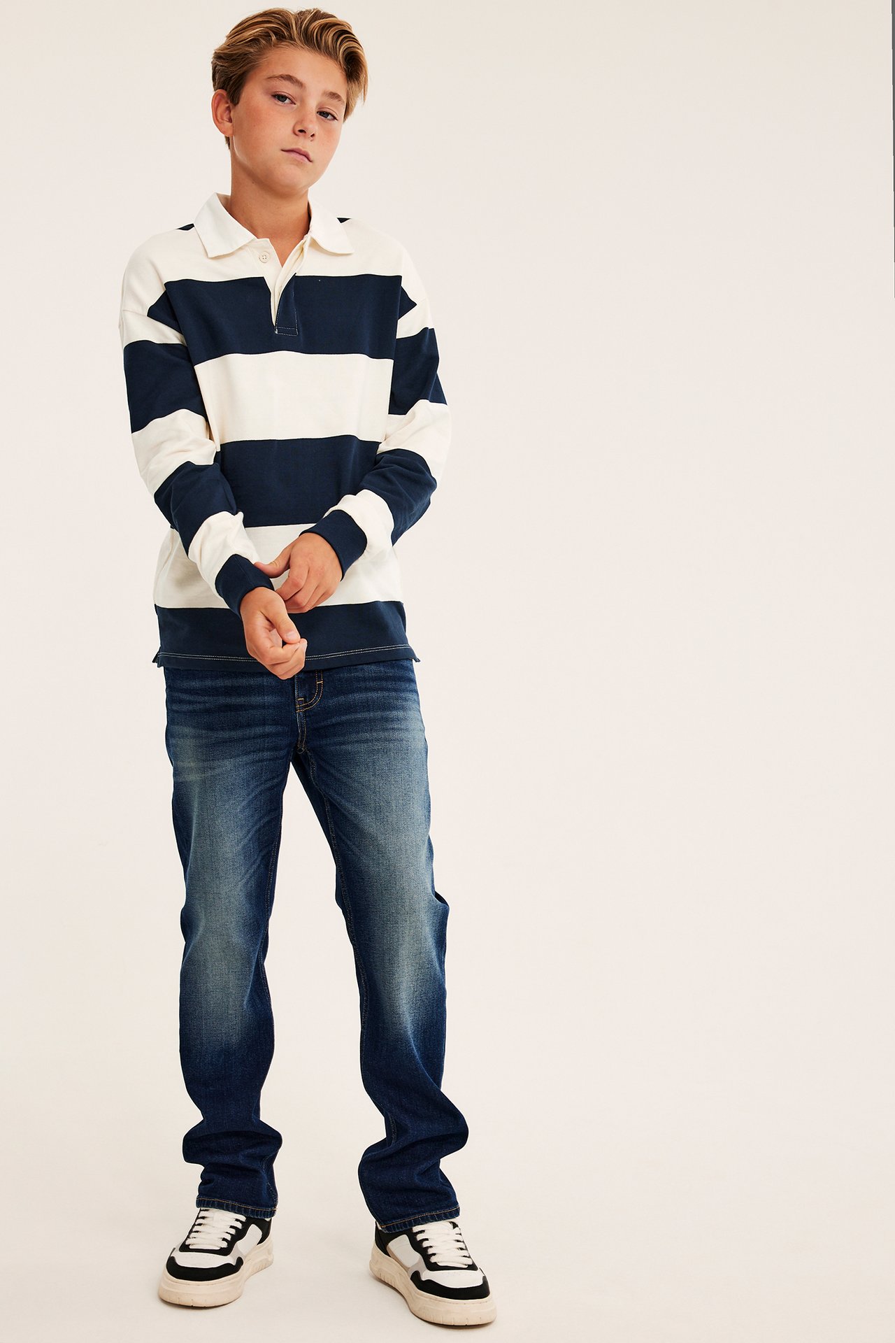 Frontbilde av en gutt i en kremhvit og marineblå stripete langermet pologenser, blå jeans og hvite joggesko.