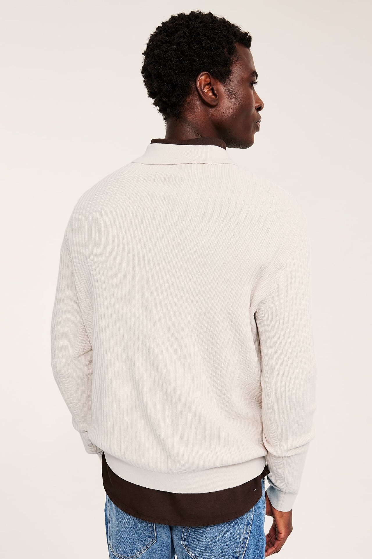 Baksidebilde av voksen mann i off-white ribbestrikket genser med krage, brun skjorte, blå jeans.