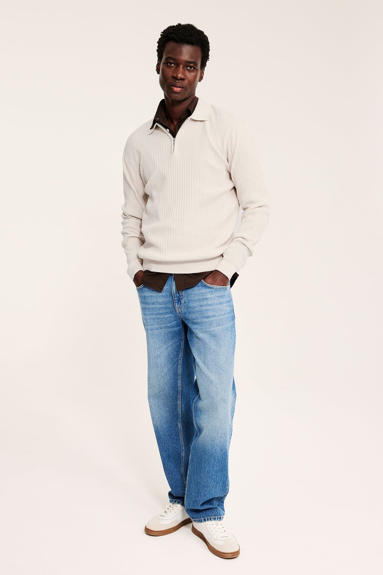 Forfra mann i beige ribbestrikket halv glidelås-genser over brun skjorte, blå jeans og hvite sneakers.