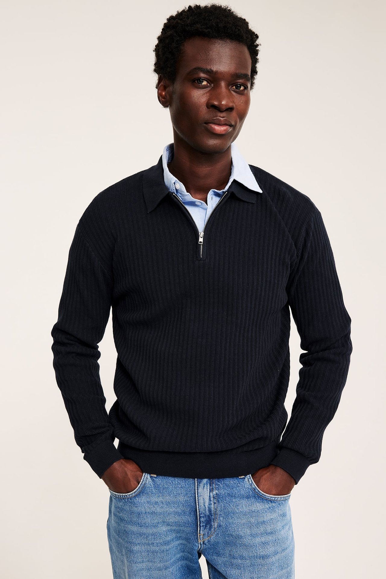 Herregenser i mørkeblå ribbestrikk med halv glidelås over lyseblå skjorte og lyseblå jeans.