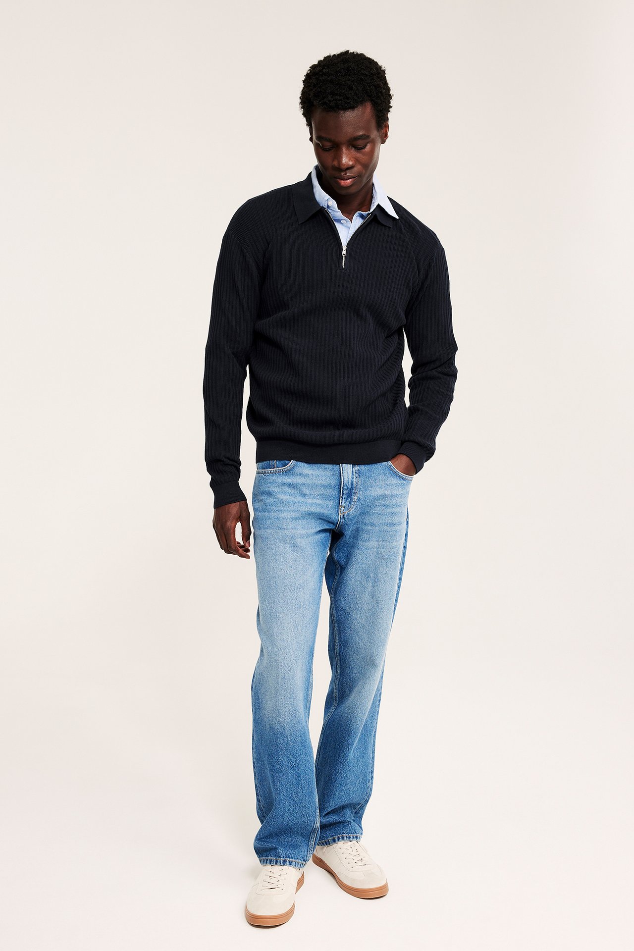 Frontbilde av mann i svart ribbet halvzip-polo, blå skjorte, blå jeans og beige joggesko.