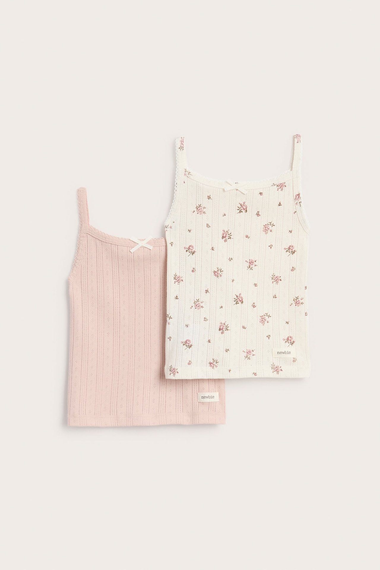 Frontvy: två ribbade baby-linnen med rosetter, ett naturvitt blommigt, ett ljusrosa.