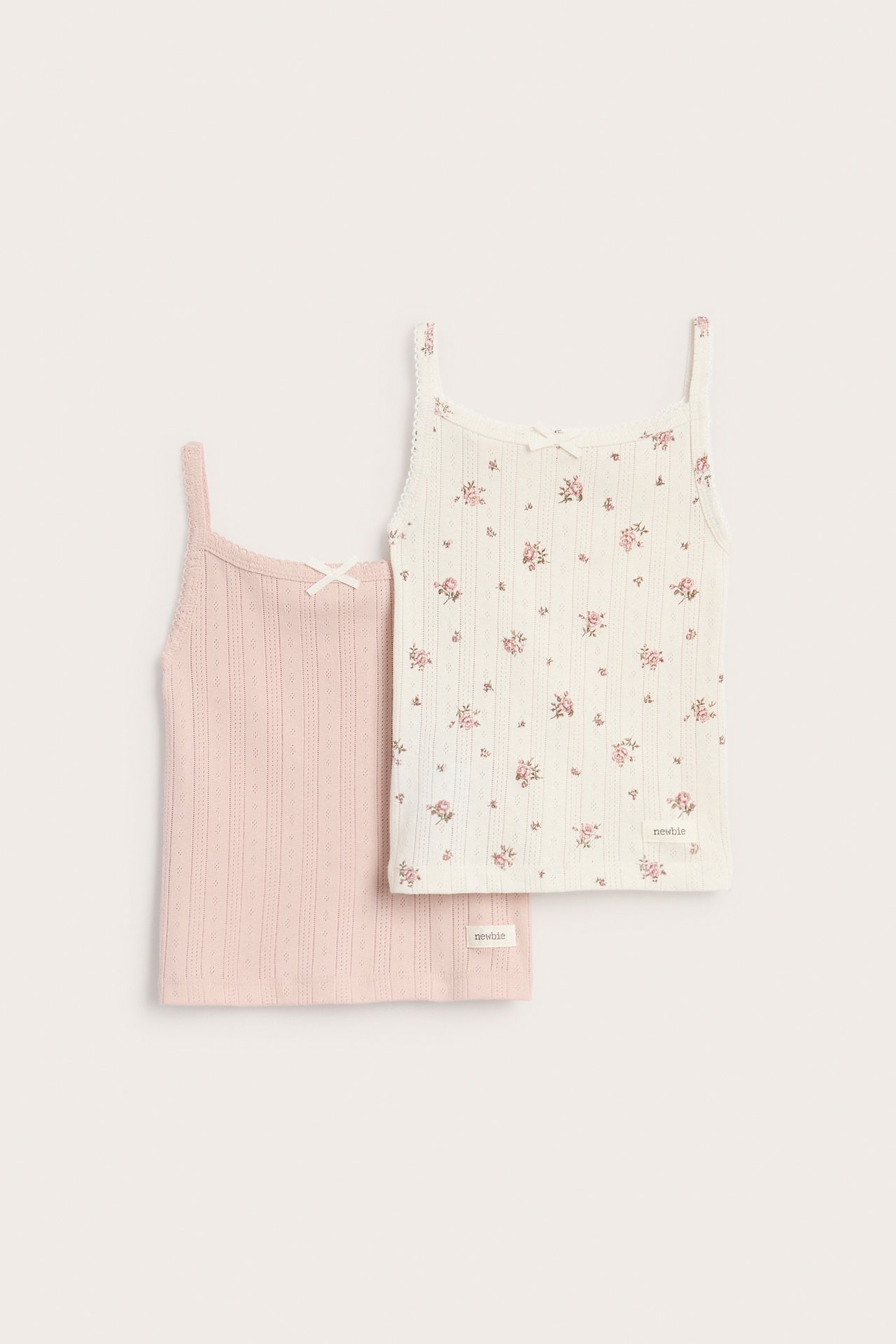 Frontvisning: to ribbestrikkede baby-singlets med sløyfer, en naturhvit blomstret, en lys rosa.