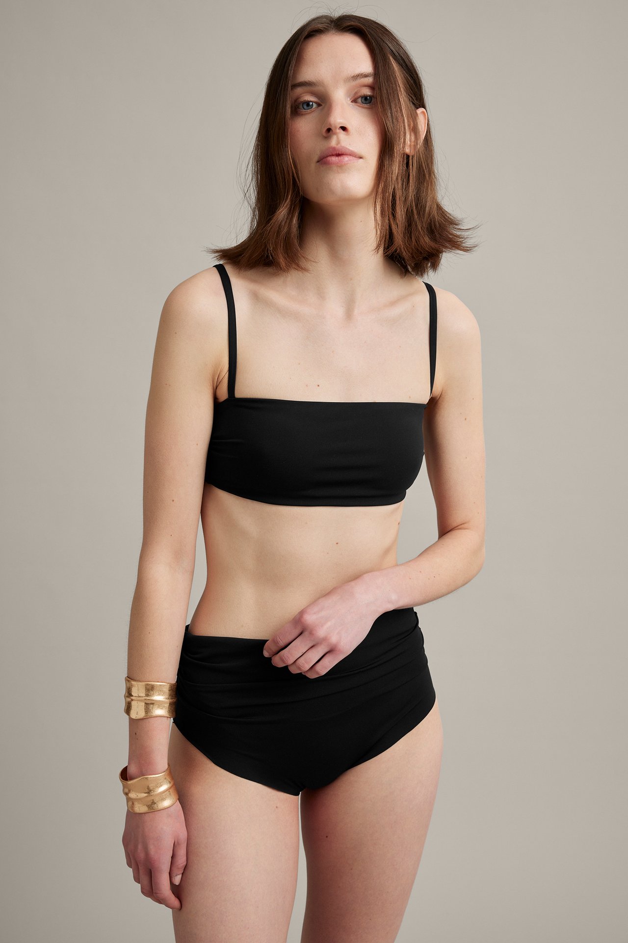 Edestä: Nainen mustassa topatussa bandeau-bikiniyläosassa, korkeavyötäröisissä alaosissa ja kultaisissa rannekoruissa.