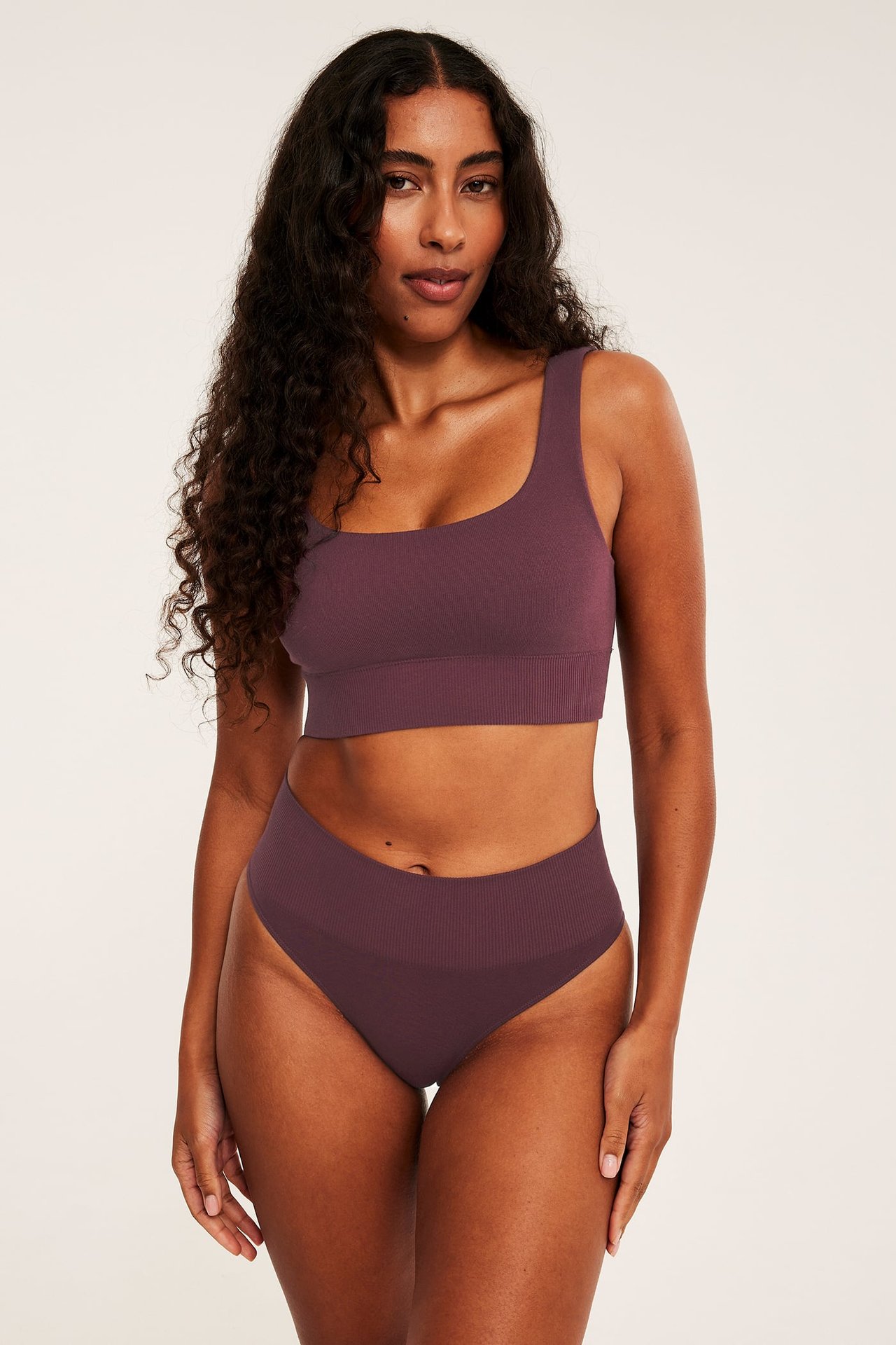 Frontvy av en kvinnlig modell i plommonfärgad ribbad sömlös bralette och matchande högmidjade trosor.