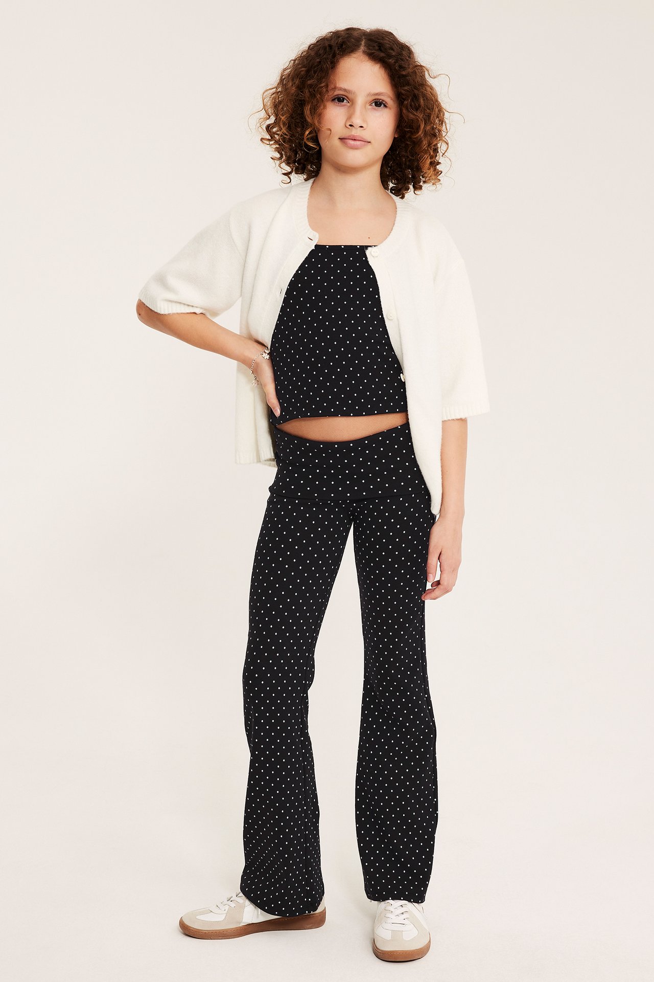 Dziecko z przodu: czarne kropkowane spodnie do jogi, pasujący top, jasny kardigan i białe sneakersy.