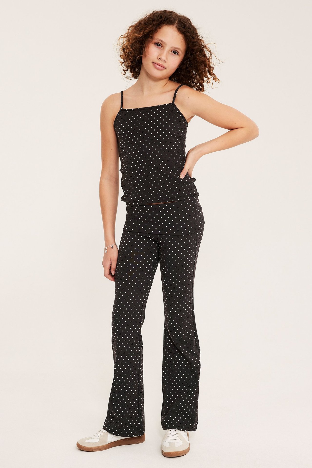 Framifrån, ett barn i svart prickigt linne, matchande utsvängda byxor och sneakers.