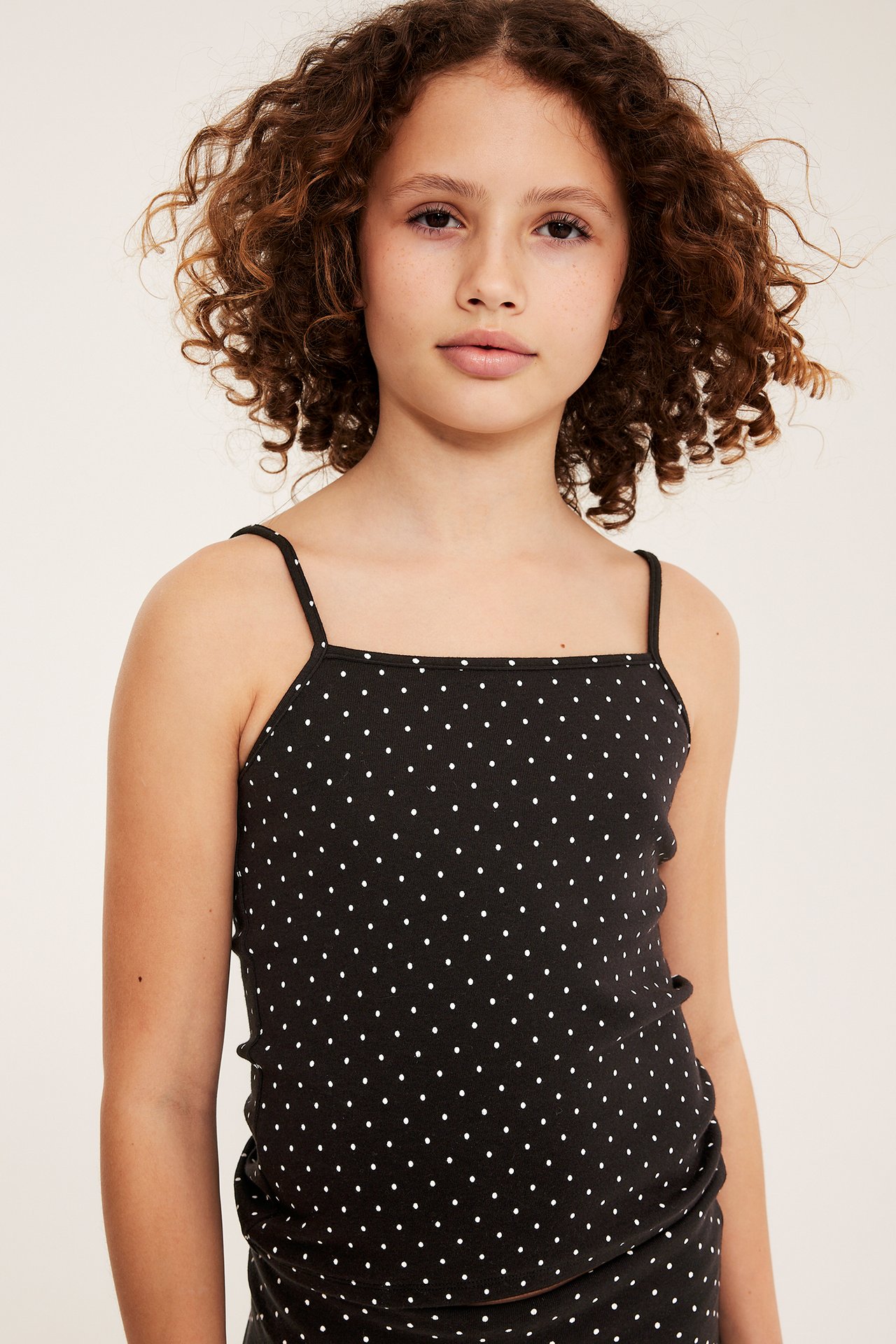 Framifrån, ett barn i svart prickigt linne i bomullstrikå med smala axelband och matchande underdel.