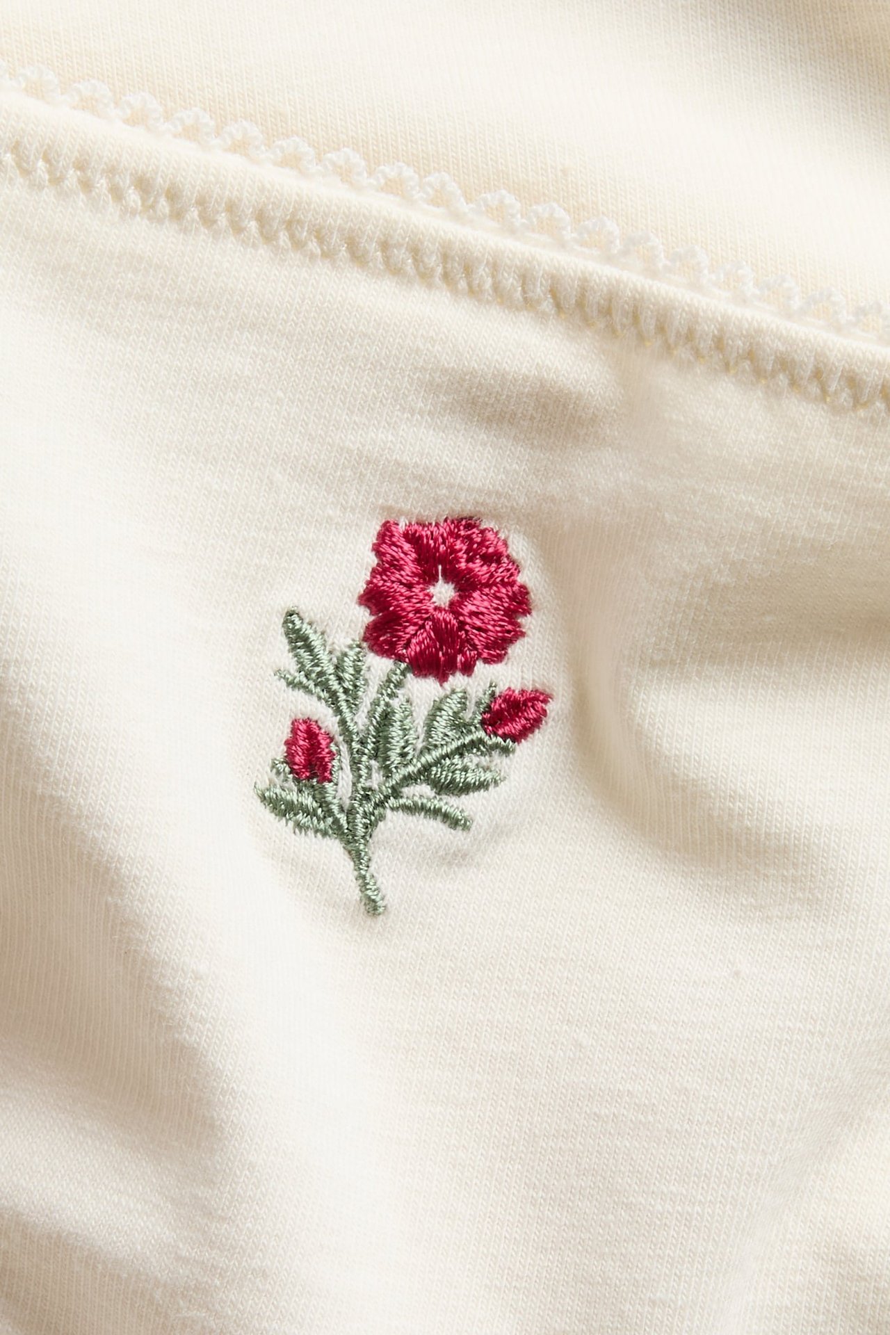 Nærbilde av offwhite bomulls-Brazilian-truse med en rød brodert blomst og bølget kant.