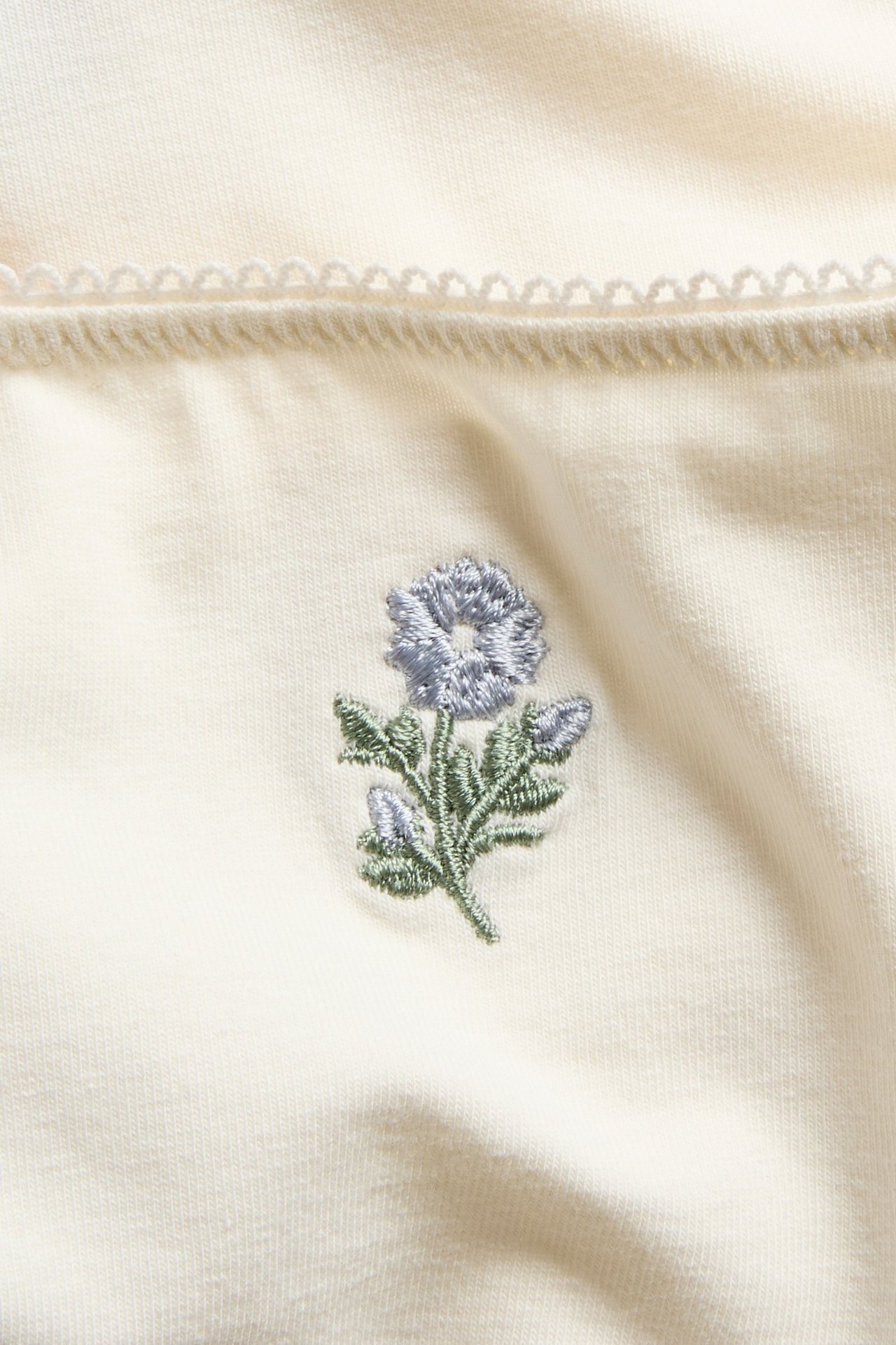Nærbilde: Offwhite bomulls-Brazilian truse med blå brodert blomst og bølget kant.