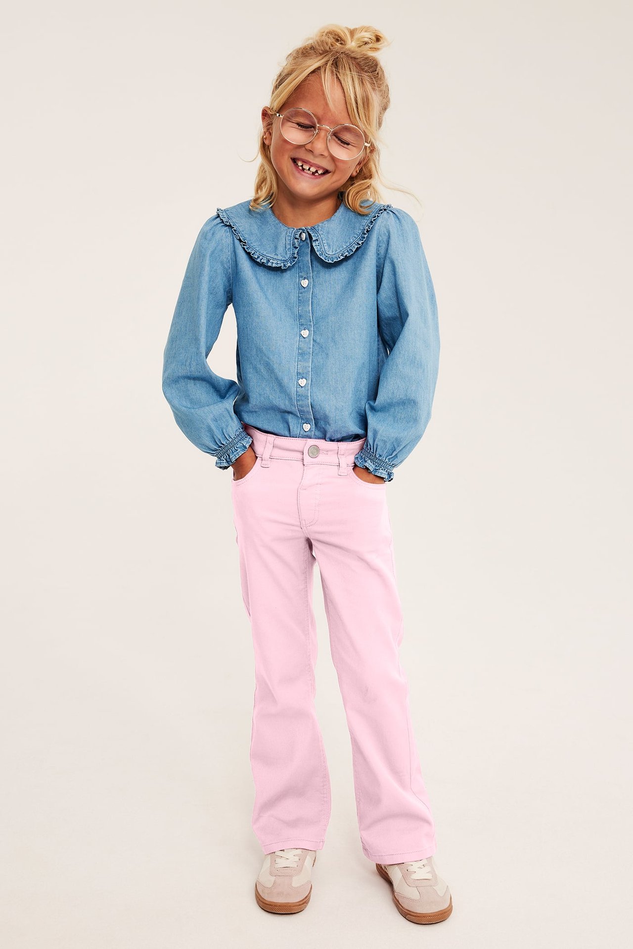 Front view: Kid wears rosa utsvängda jeans, en blå jeansskjorta med volangkrage och beiga sneakers.