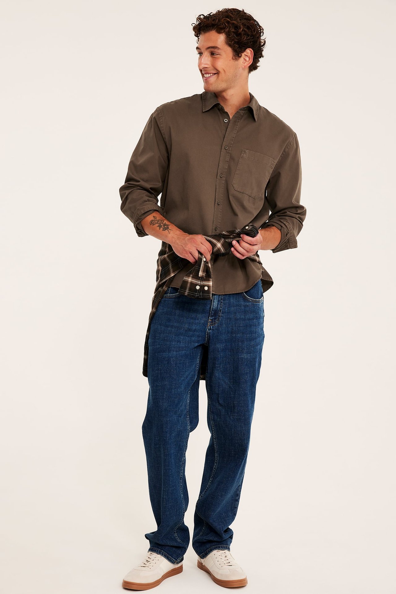 Frontvy: olivgrön långärmad skjorta, mörkblå jeans, rutig skjorta knuten i midjan och beige sneakers.
