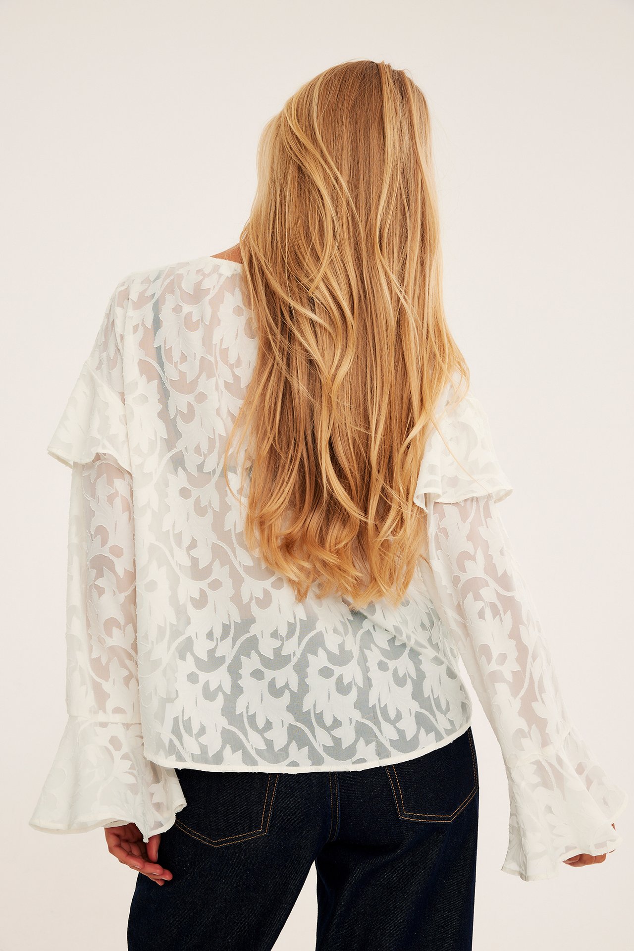 Kvinnlig modell, bakifrån, bär en vit skir mönstrad blus med volanger och mörkblå jeans.