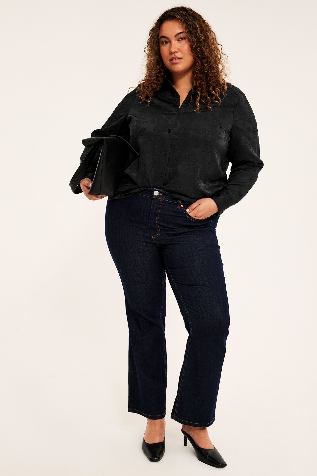 Frontbilde av kvinne i svart mønstret skjorte, mørkeblå jeans, svarte mules, holder veske.