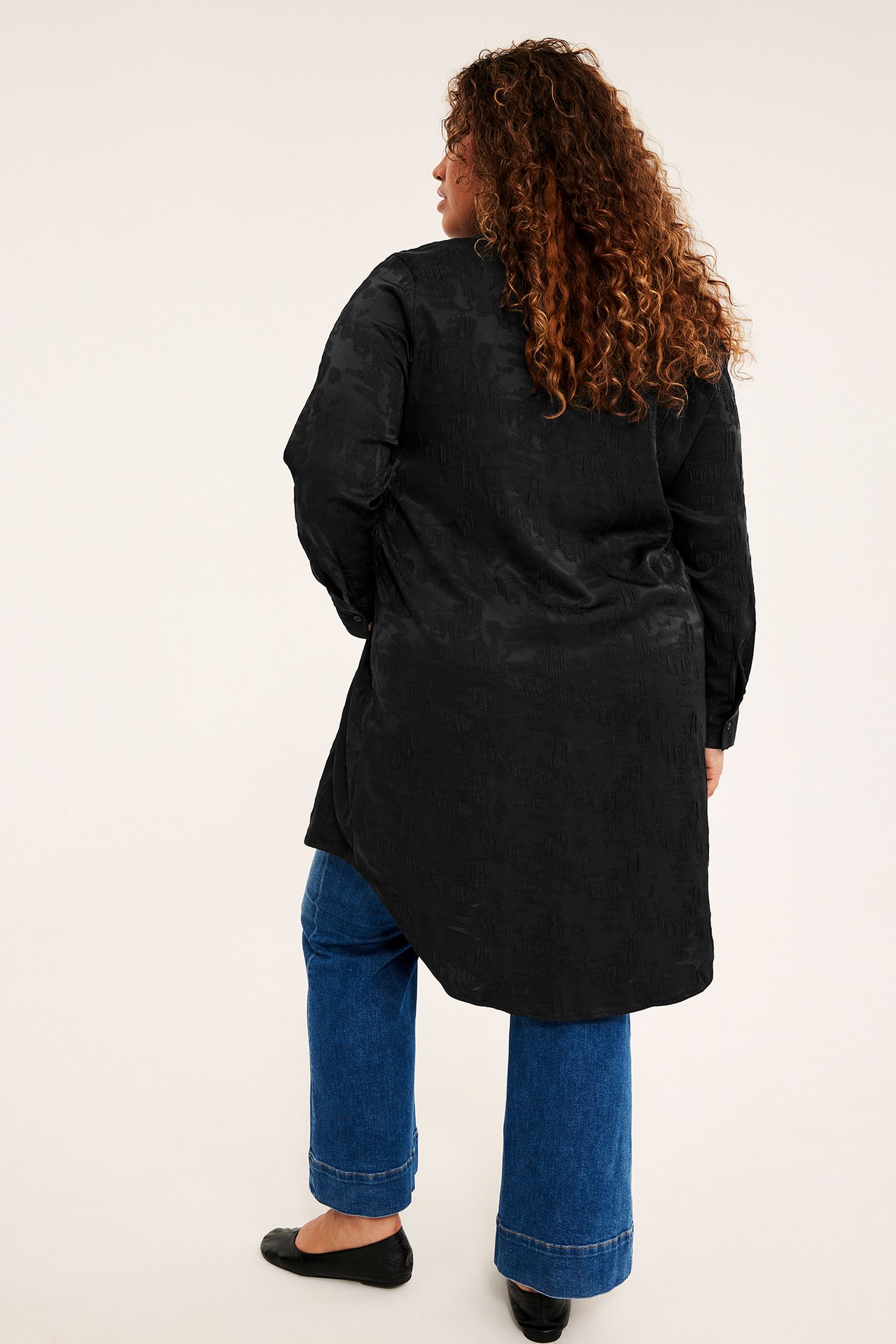 Ryggvisning av kvinnelig modell iført sort teksturert lang tunika, blå jeans og svarte flate sko.