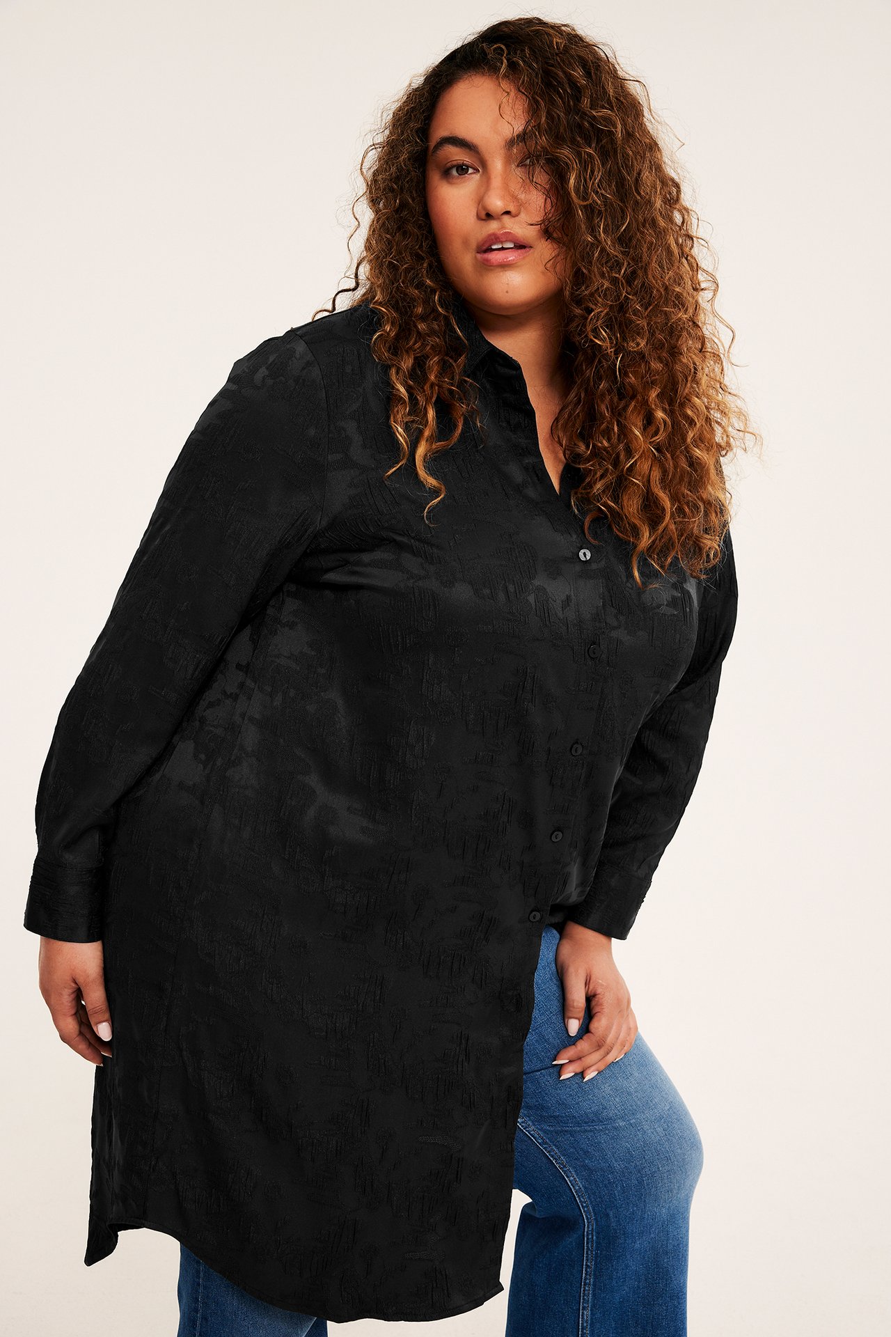Forfra, kvinnelig modell i en lang svart teksturert tunikaskjorte og blå jeans.