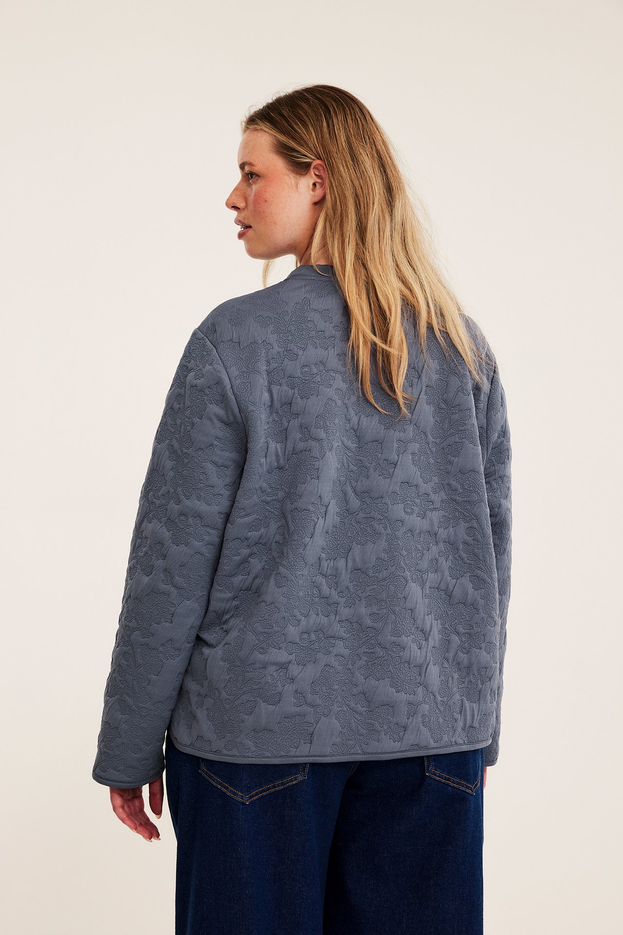 Kvinna bakifrån i blågrå kviltad blommig jacka och mörkblå jeans.
