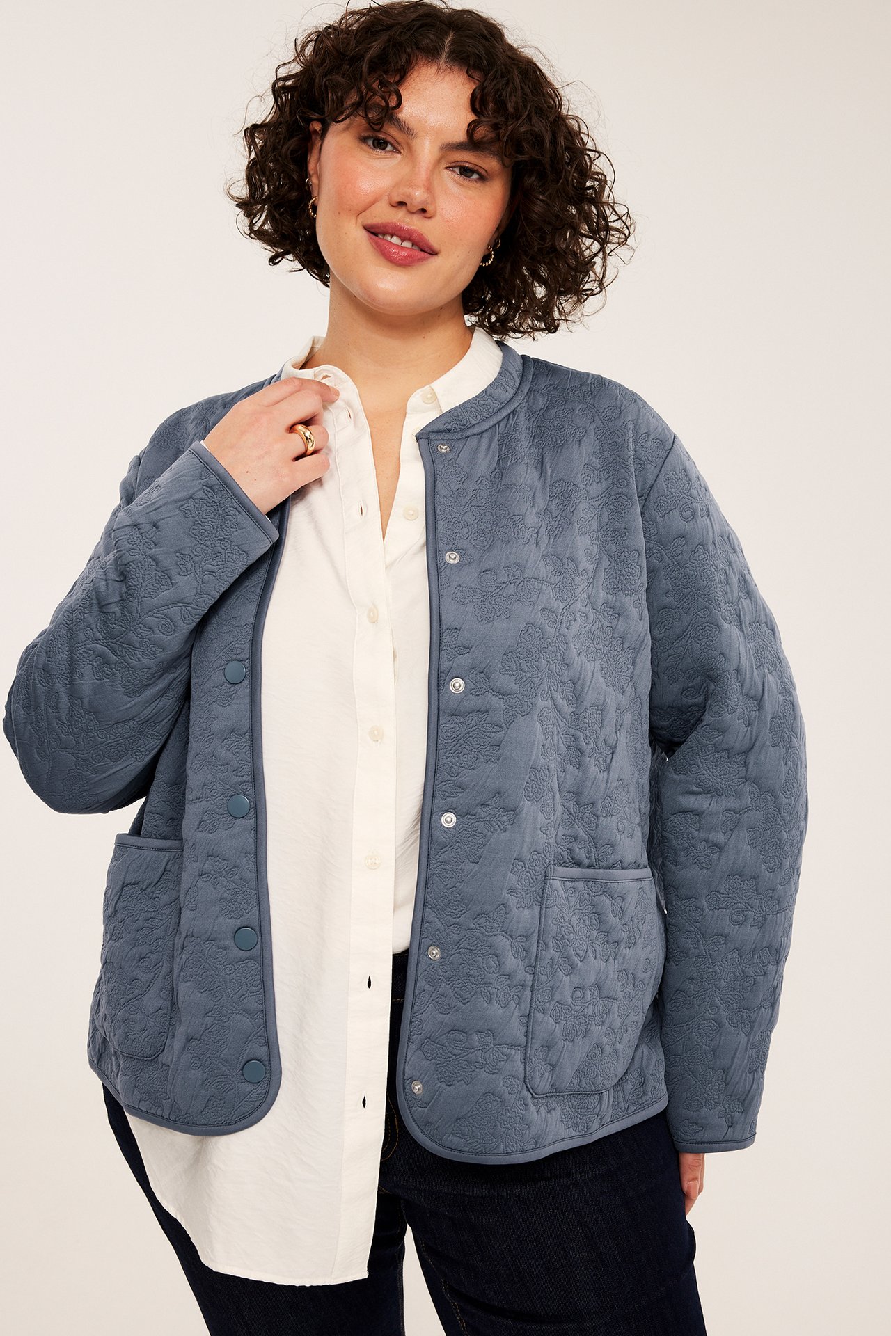 Framifrån, kvinna i blå quiltad blommig jacka över vit skjorta och mörka jeans.