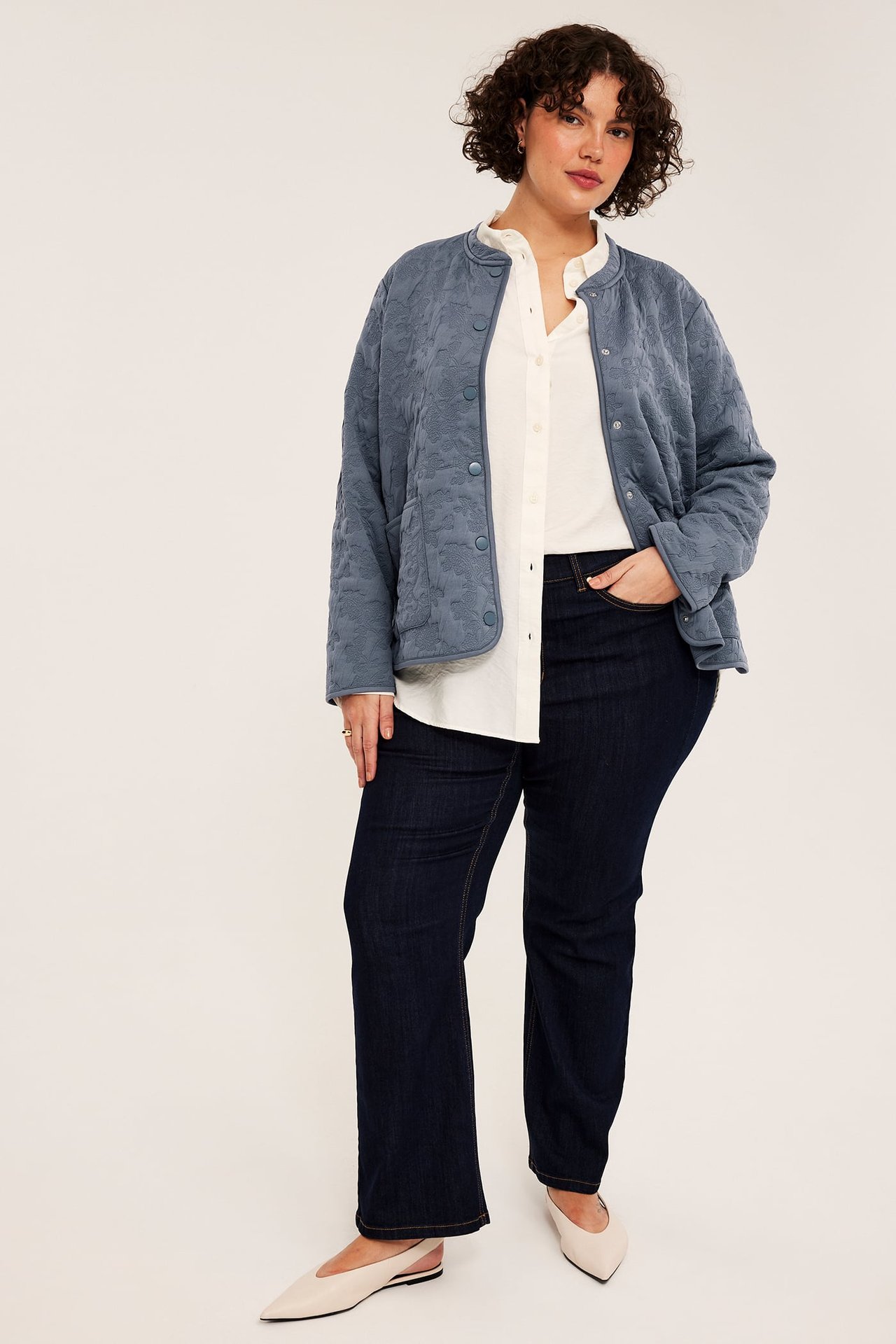 Framsida av kvinnlig modell i blå quiltad jacka, vit skjorta, mörkblå jeans och gräddfärgade flats.
