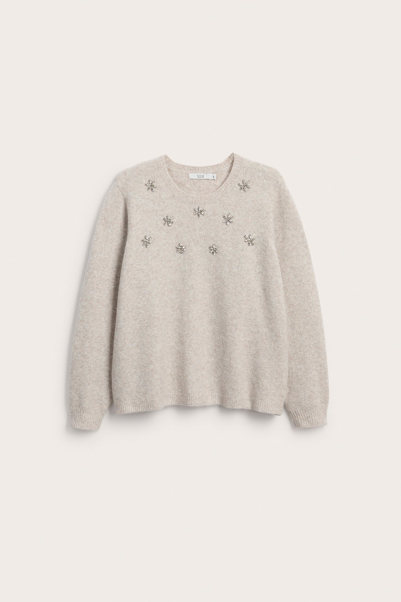 Framsida av en ljusbeige tröja med rund hals och silverfärgade utsmyckningar.