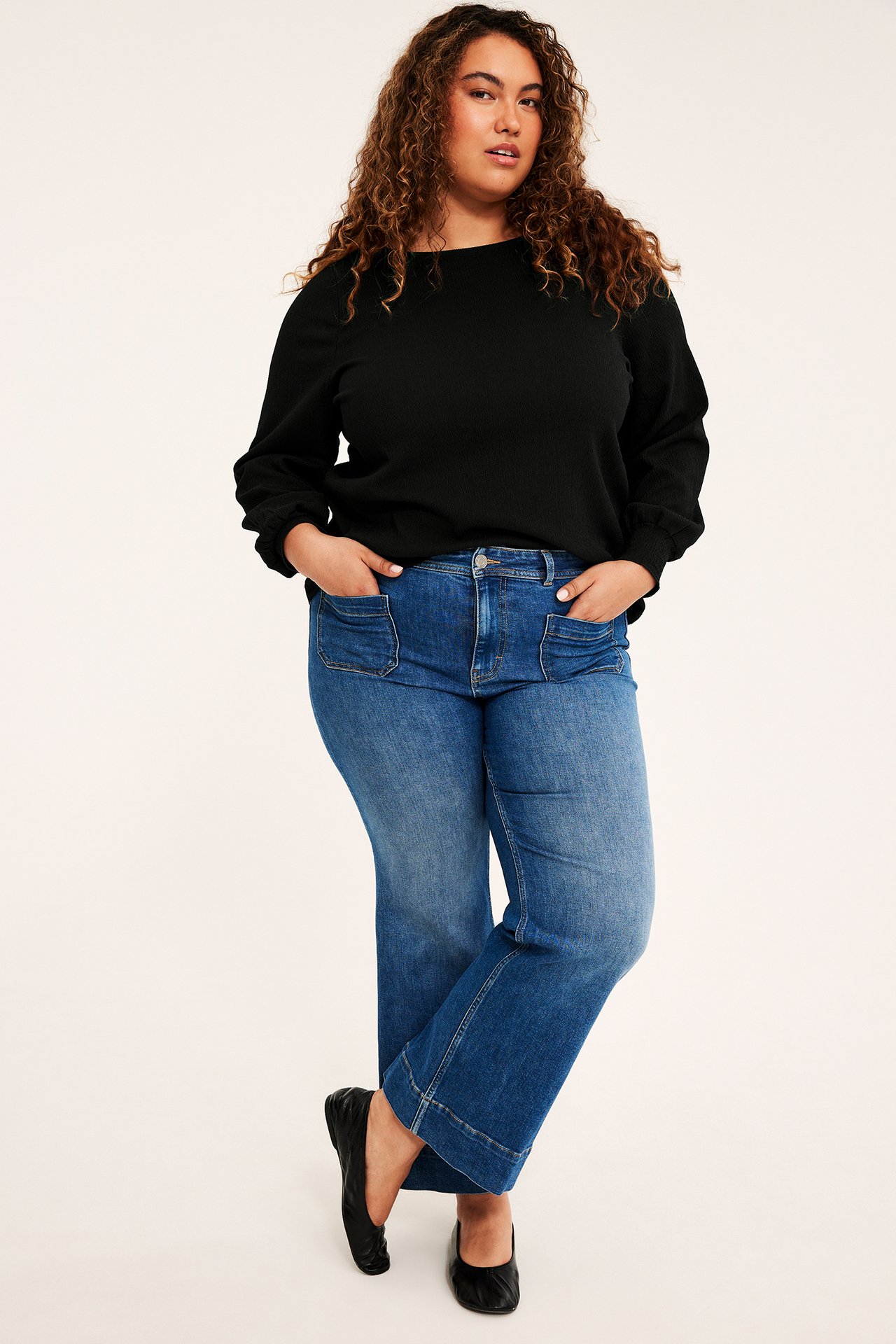 Framifrån: Kvinna i blå vida jeans med framfickor, svart topp och ballerinaskor.
