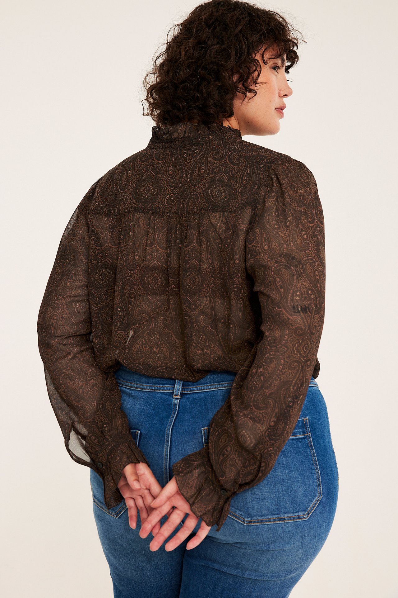 Kvinne bakfra i brun, gjennomsiktig paisley-bluse og blå jeans.