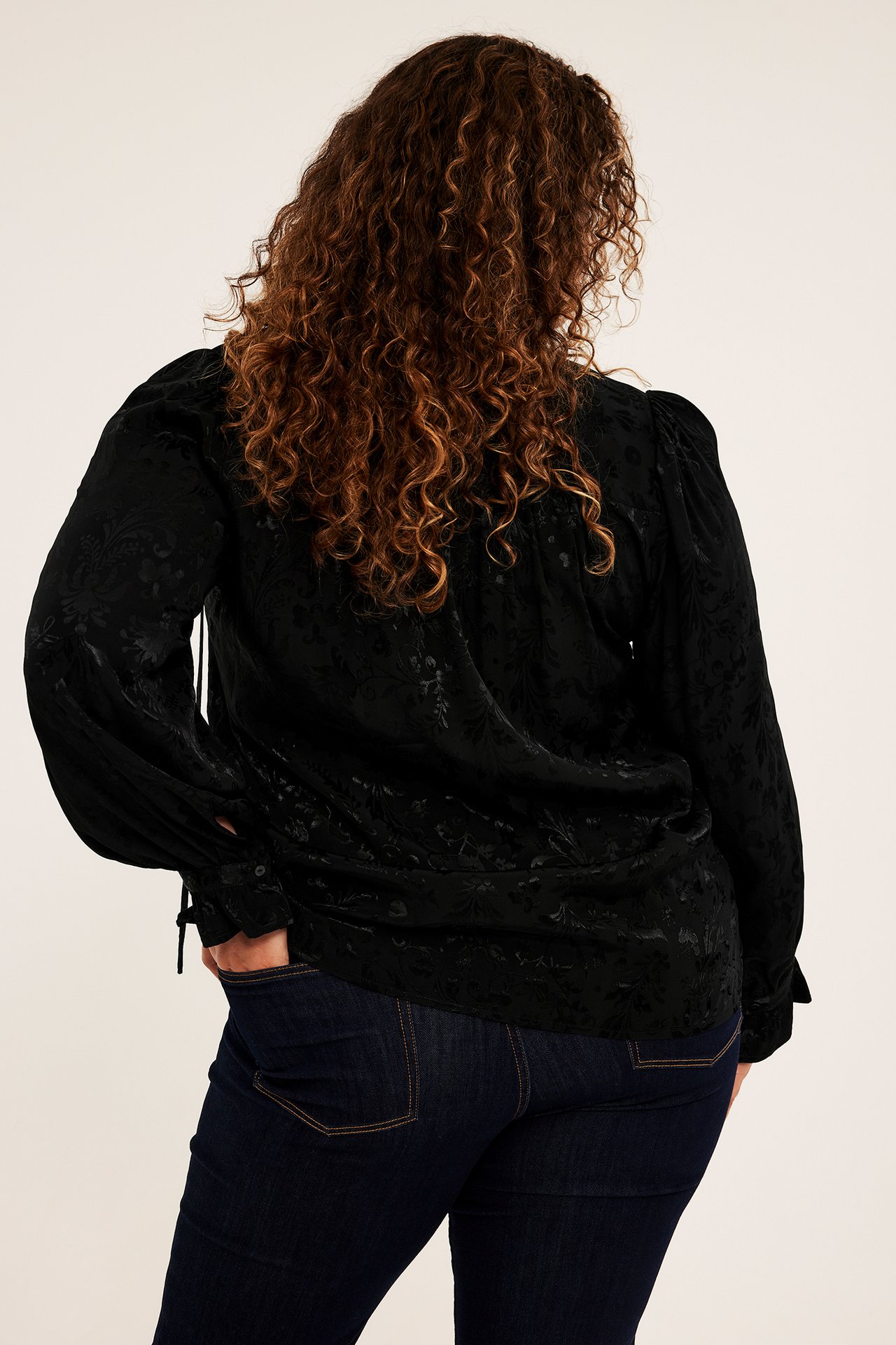 Kvinnelig modell bakfra i svart mønstret langermet bluse med puffermer og mørke jeans.