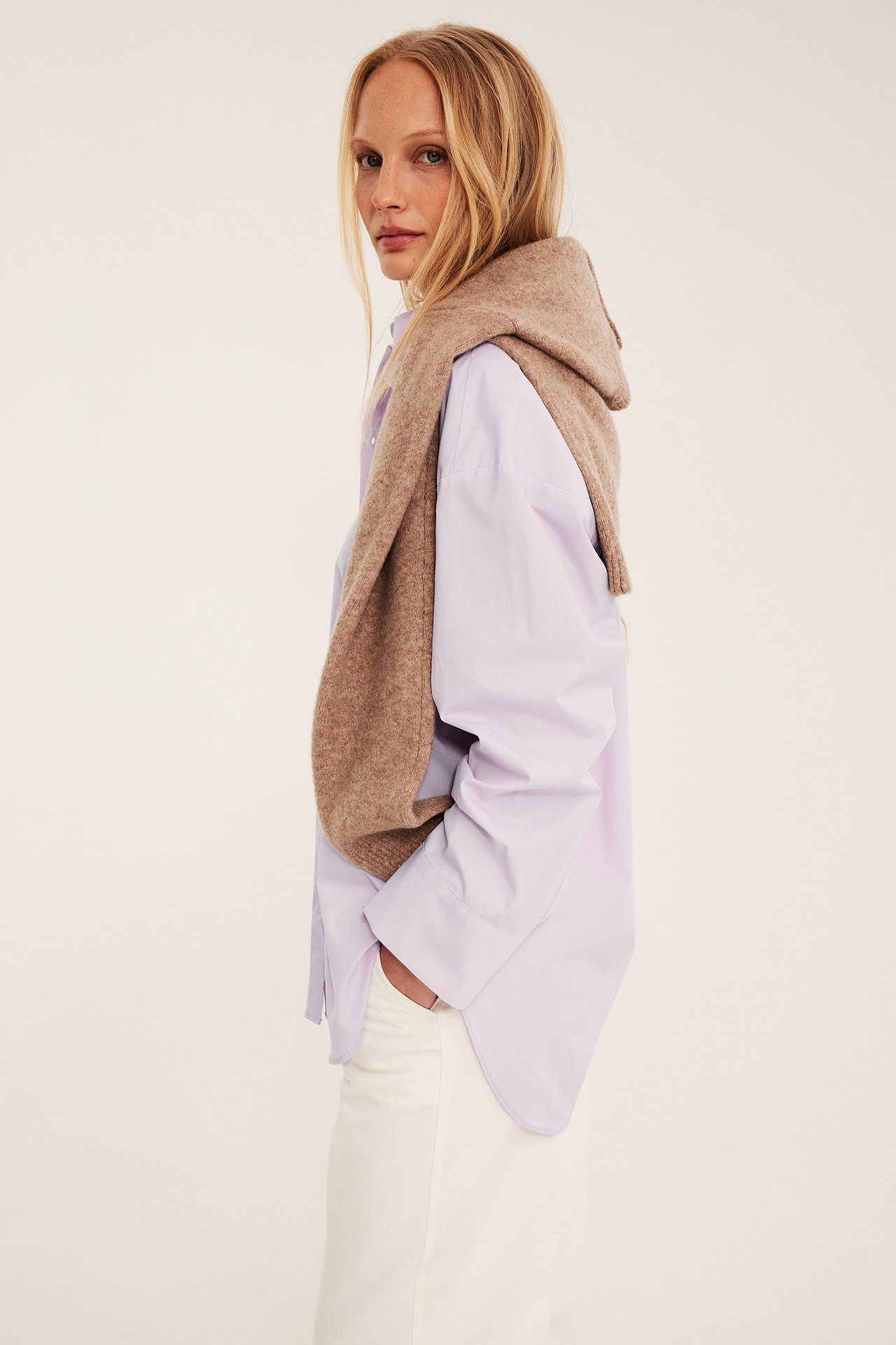 Sidovy av kvinna i ljuslila oversized bomullsskjorta, med brun stickad tröja draperad och vita byxor.