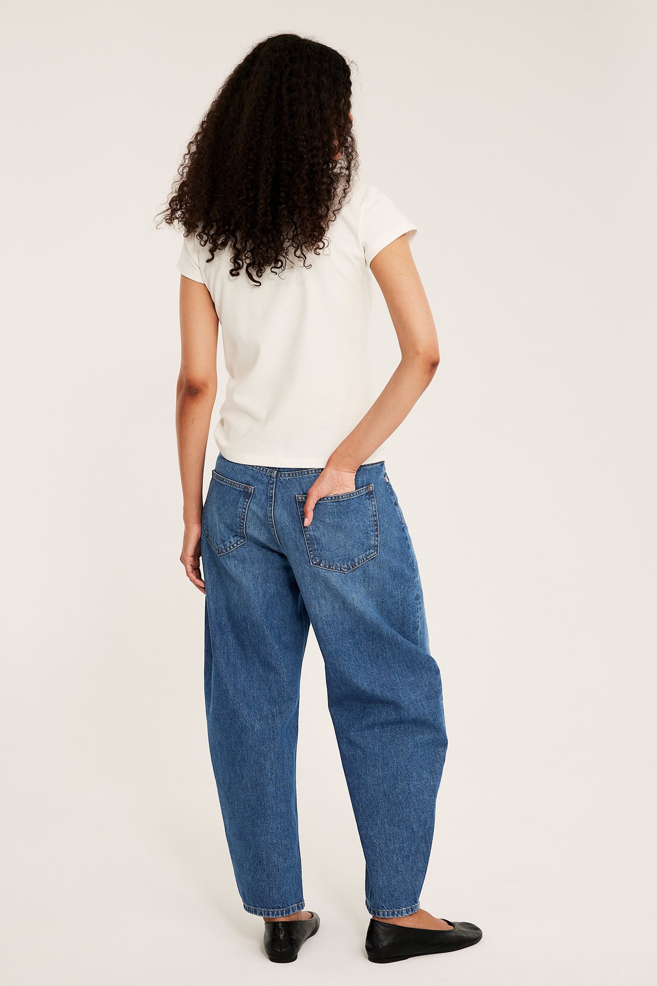 Ryggvy: kvinna i blå denim barrel jeans, hand i fickan, vit t-shirt, svarta ballerinaskor.