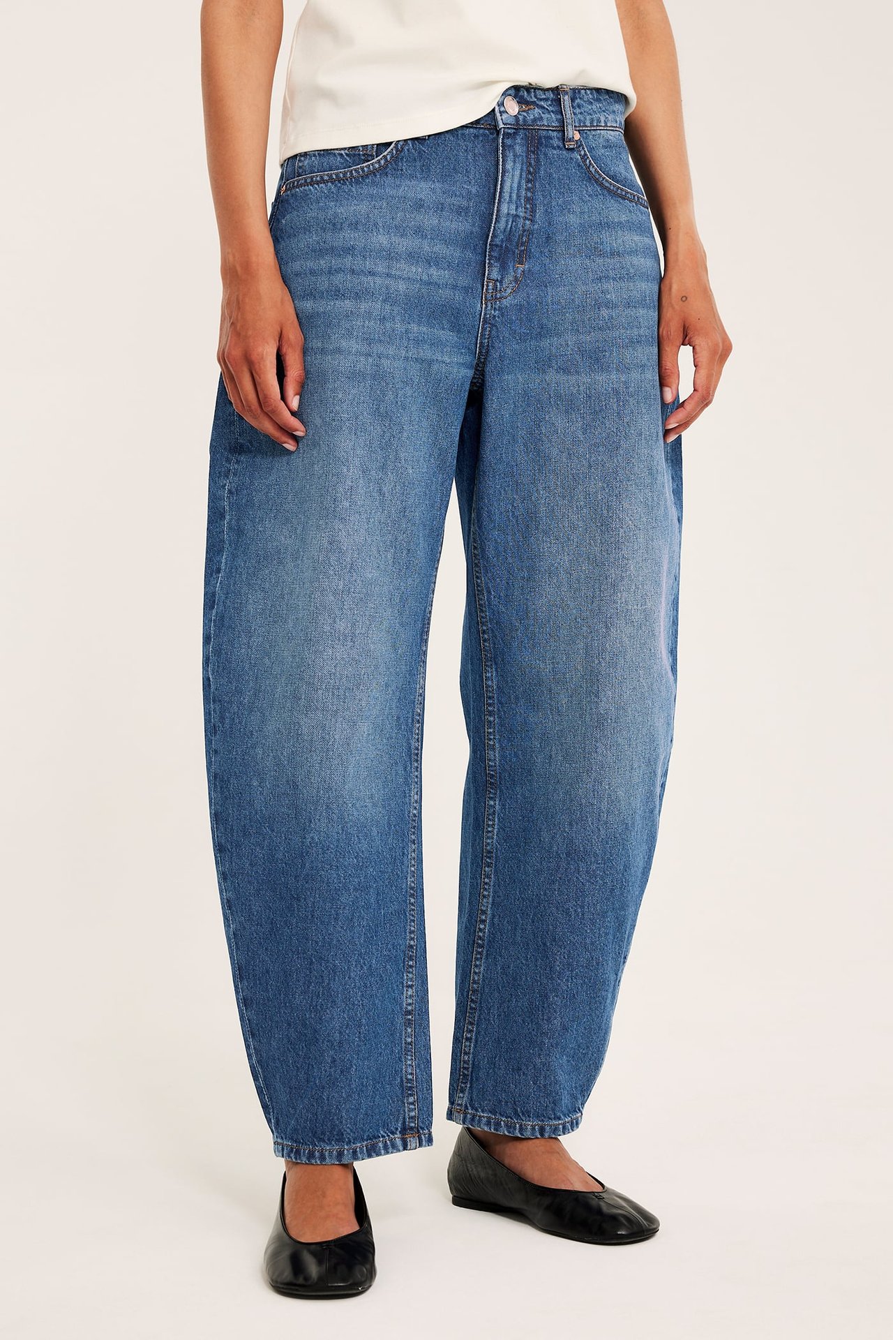 Forfra, kvinne i blå denim barrel jeans med høyt liv, hvit t-skjorte og svarte flate sko.