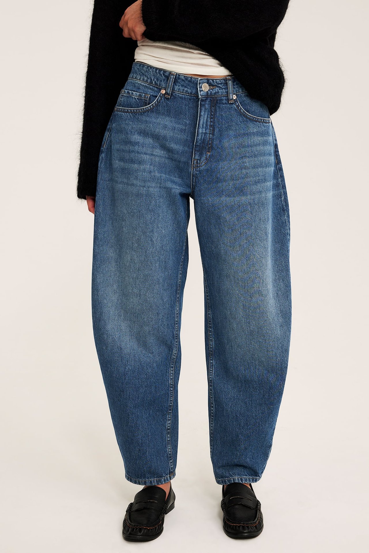 Framifrån av vuxen i lösa mellanblå denimjeans, svart tröja och svarta loafers.