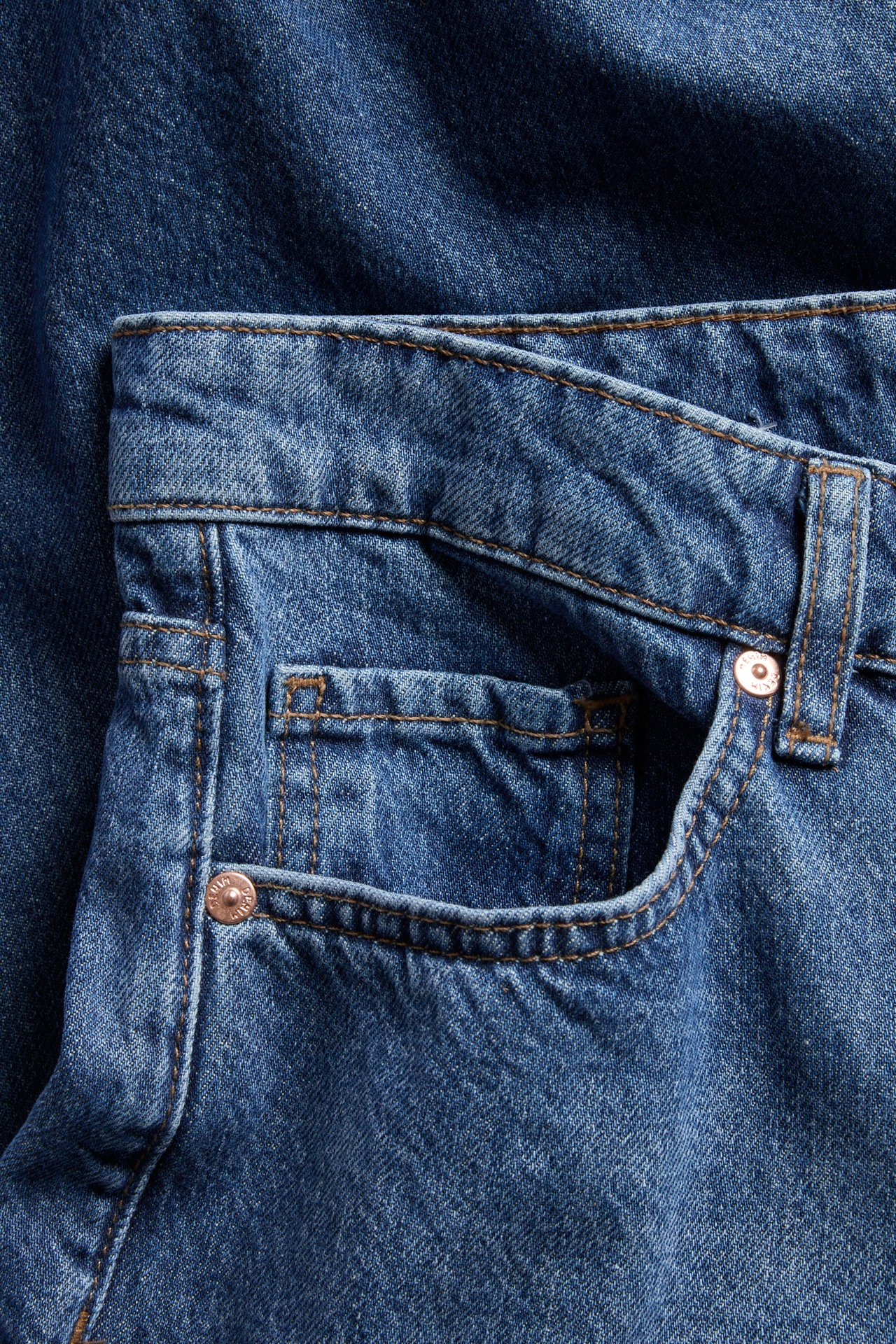 Nærbilde av blå bomullsdenim high waist-jeans med frontlomme, brune sømmer og kobbernagler.