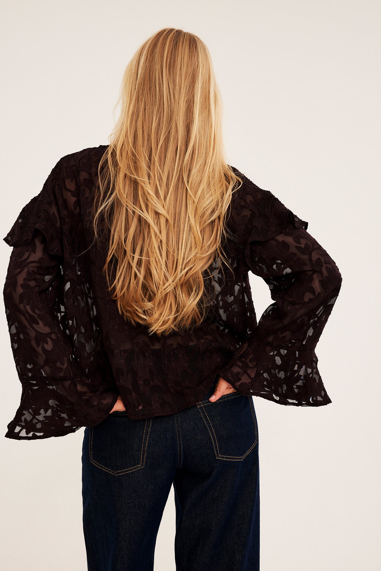Bakifrån av en kvinna i en mörkbrun skir mönstrad blus med volanger och mörkblå vida jeans.
