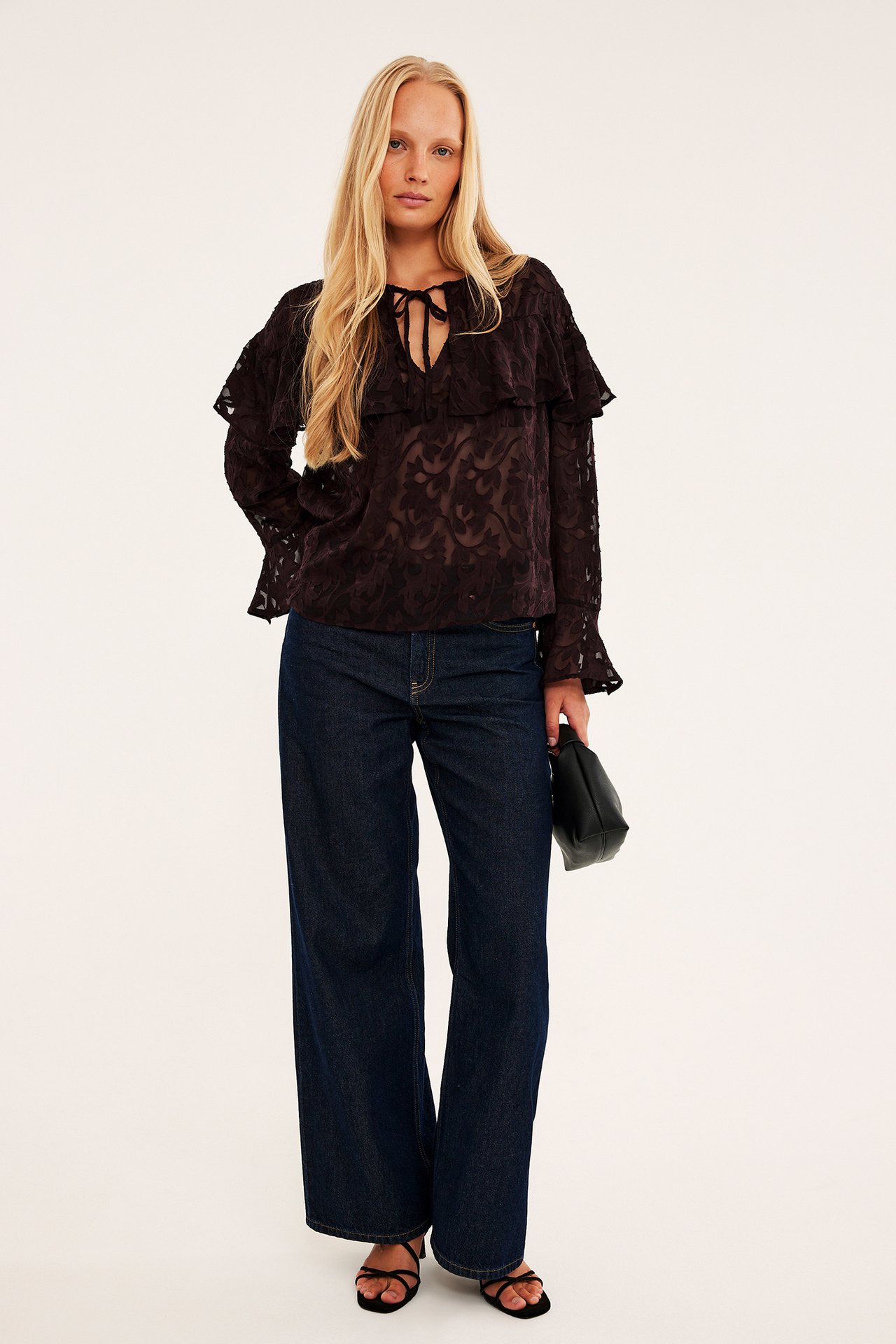 Kvinnlig modell i mörk volangblus med knyt, vida jeans, svarta sandaler och en svart clutch.