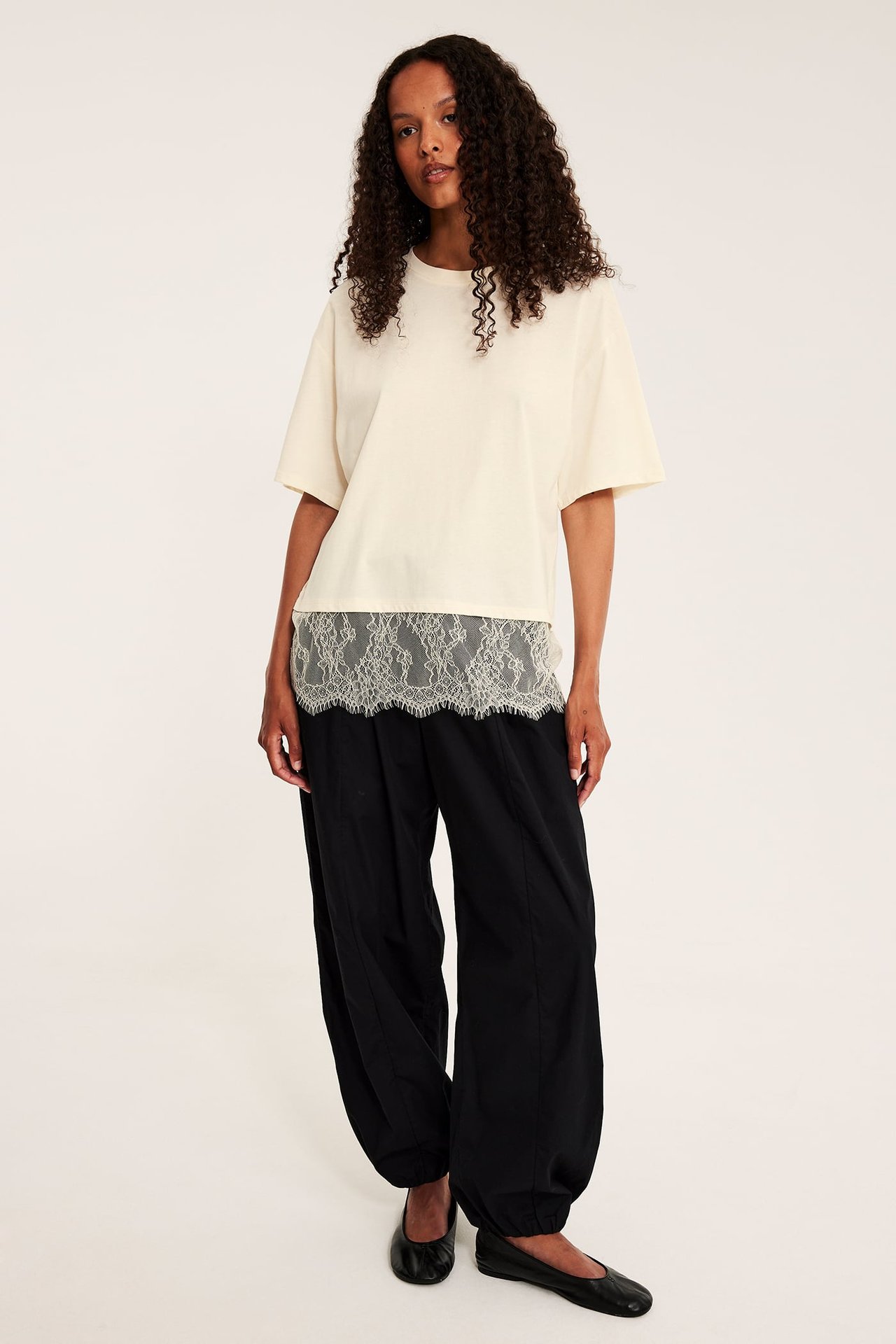 Front view av en kvinne i en off-white t-skjorte med blondekant, sorte løse bukser og sorte flate sko.