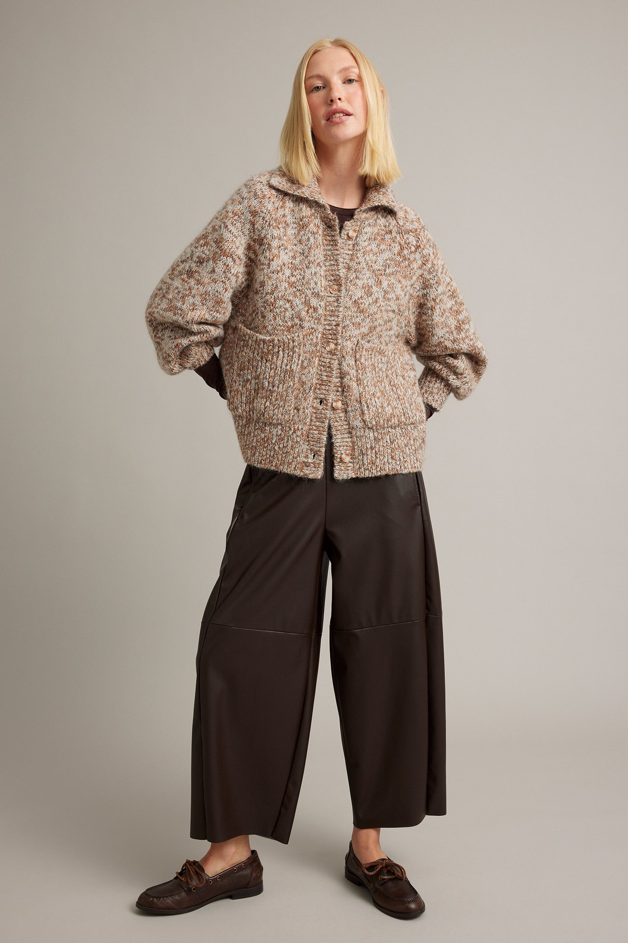 Framifrån: Kvinnlig modell i en spräcklig beige brun stickad kofta, mörkbruna vida byxor och loafers.