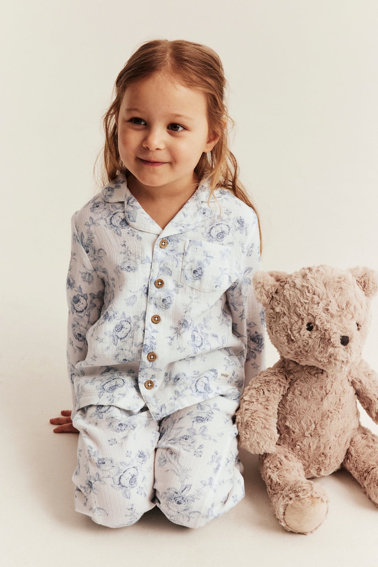 Forfra, et barn i hvit bomullsblomstrete pyjamas med knapper, ved siden av en brun teddybjørn.