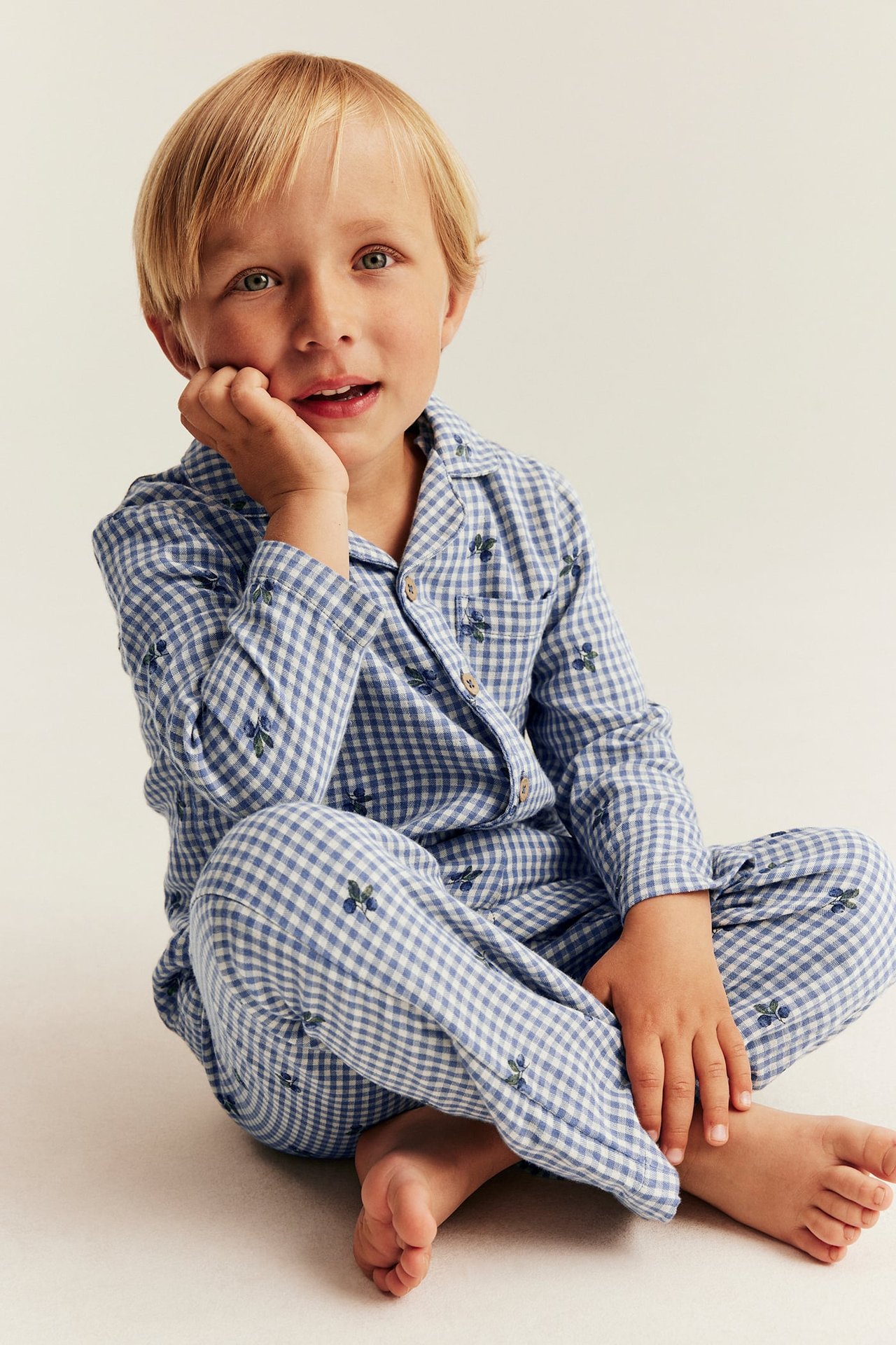 Framifrån, ett barn i blåvitrutig pyjamas med blåbärsmönster, kragad skjorta och matchande byxor.