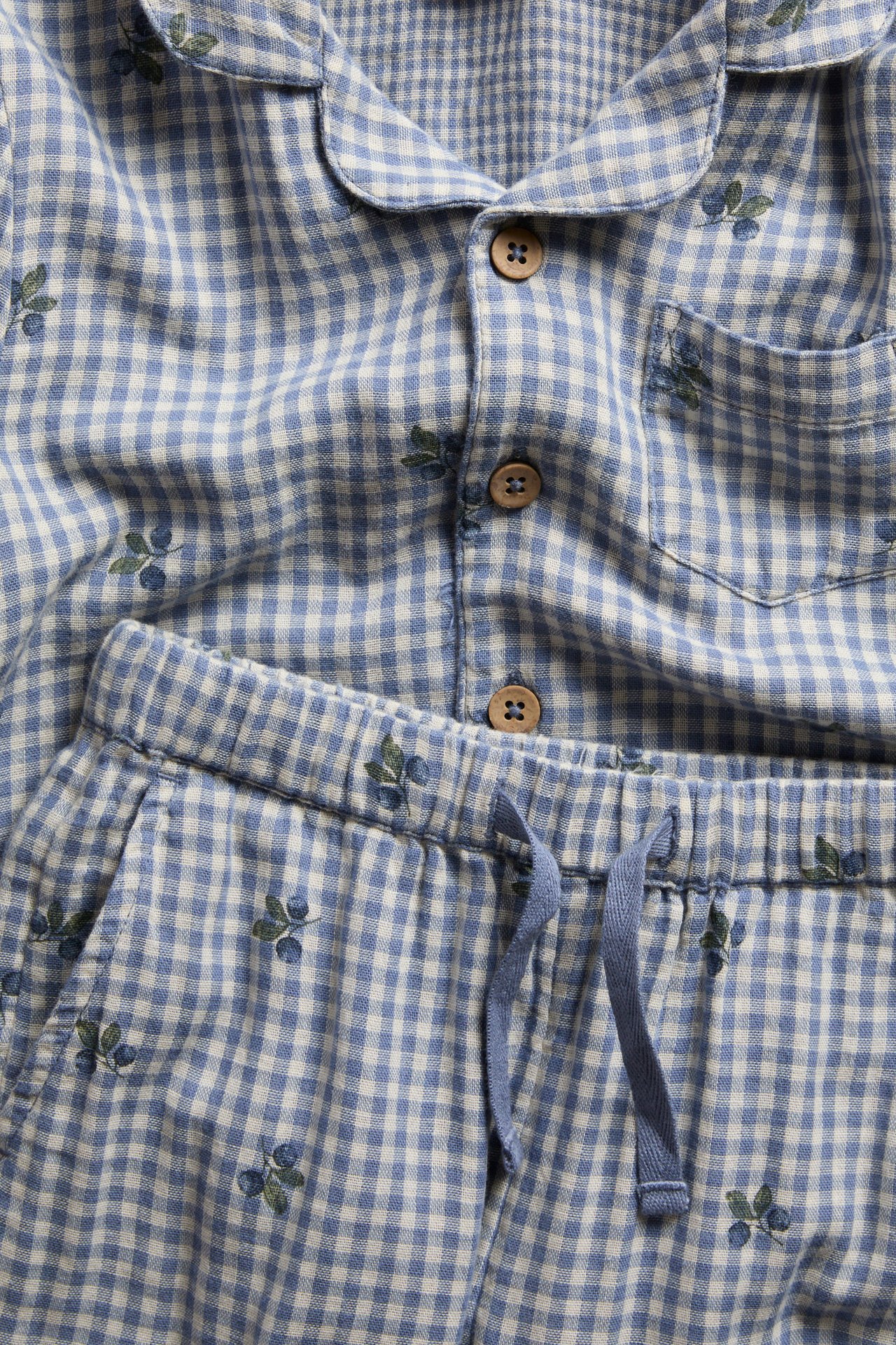 Lähikuva sinivalkoisesta ruudullisesta puuvillapyjamasta hedelmäkuviolla. Kauluspaita napeilla, housut nyörillä.