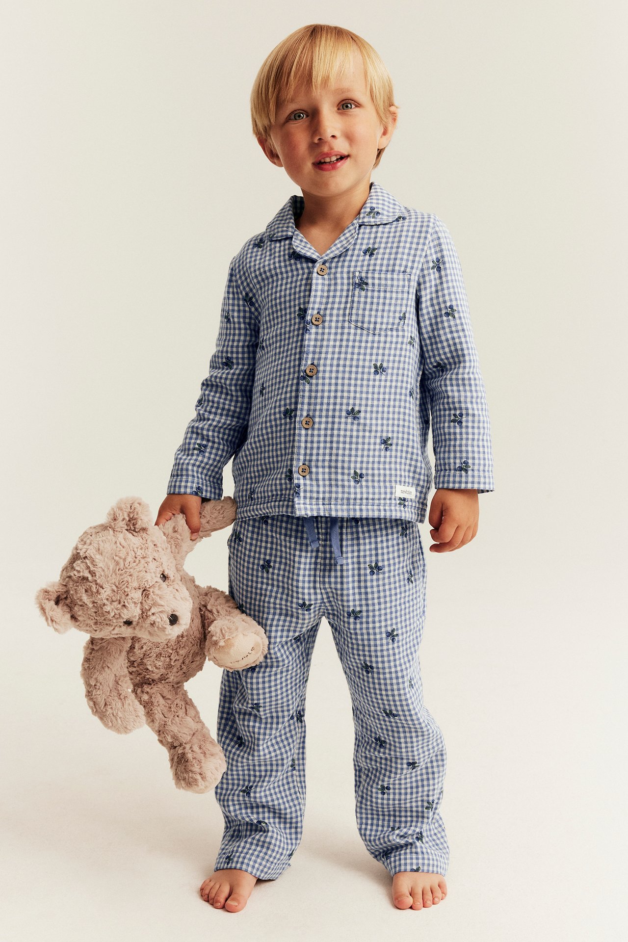 Edestä. Lapsi siniruudullisessa marjakuvioisessa pyjamassa, pitää vaaleanruskeaa nallea kädessään.