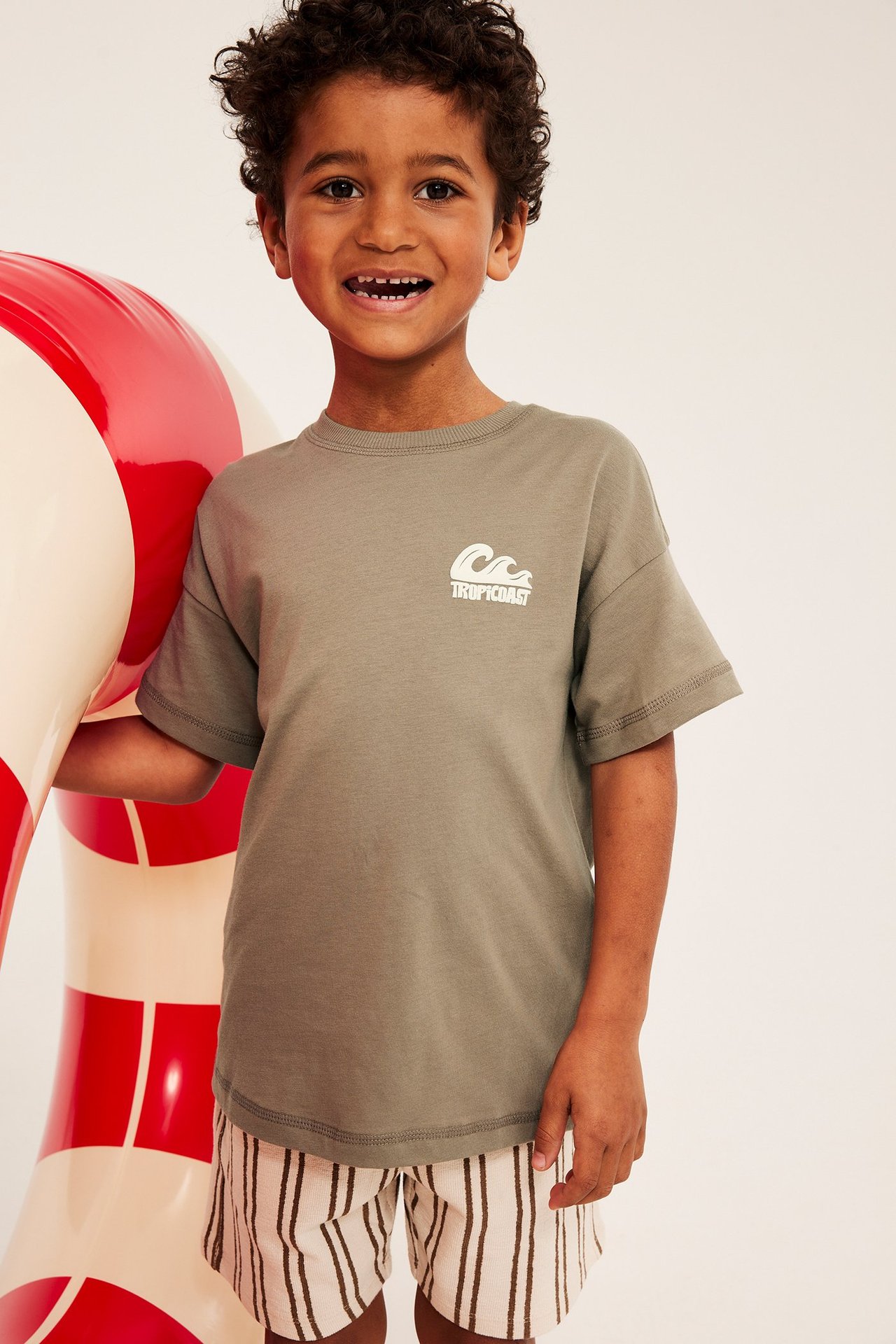 Framifrån: ett barn i brun oversize t-shirt med vitt tryck och vitrandiga shorts.