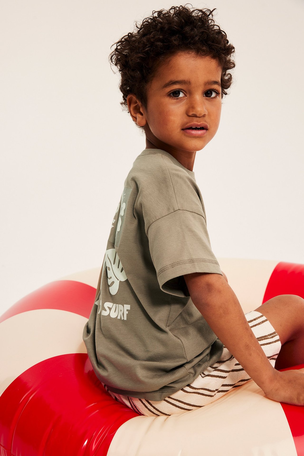 Bakifrån: Kid i brun oversize bomulls-t-shirt med ryggtryck, randiga shorts, sitter på badring.