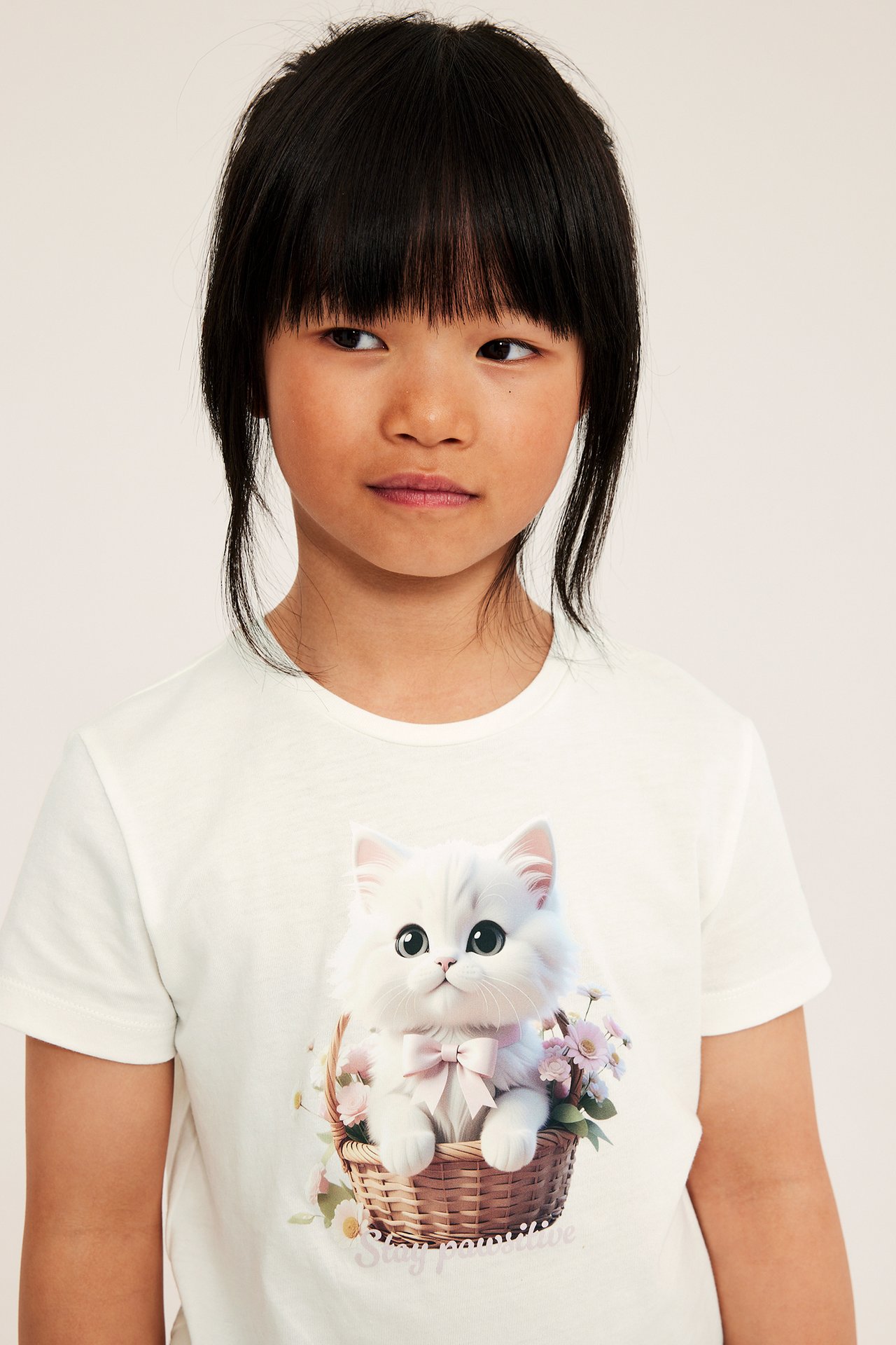 Framifrån: Ett barn bär en offwhite bomulls-T-shirt med tryck av en fluffig vit katt i korg.