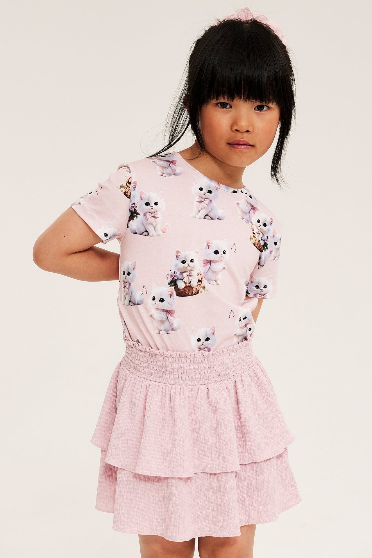 Edestä: Lapsi pukeutuu vaaleanpunaiseen puuvillaiseen kissakuvioituun T-paitaan, vaaleanpunaiseen röyhelöhameeseen ja hiuslenkkiin.