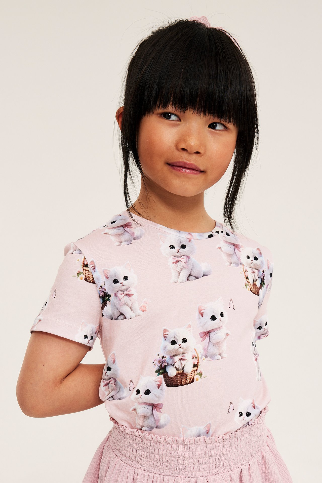 Framifrån: Ett barn bär en ljusrosa bomulls-t-shirt med kattmönster och rosa rosetter, samt en rosa kjol.