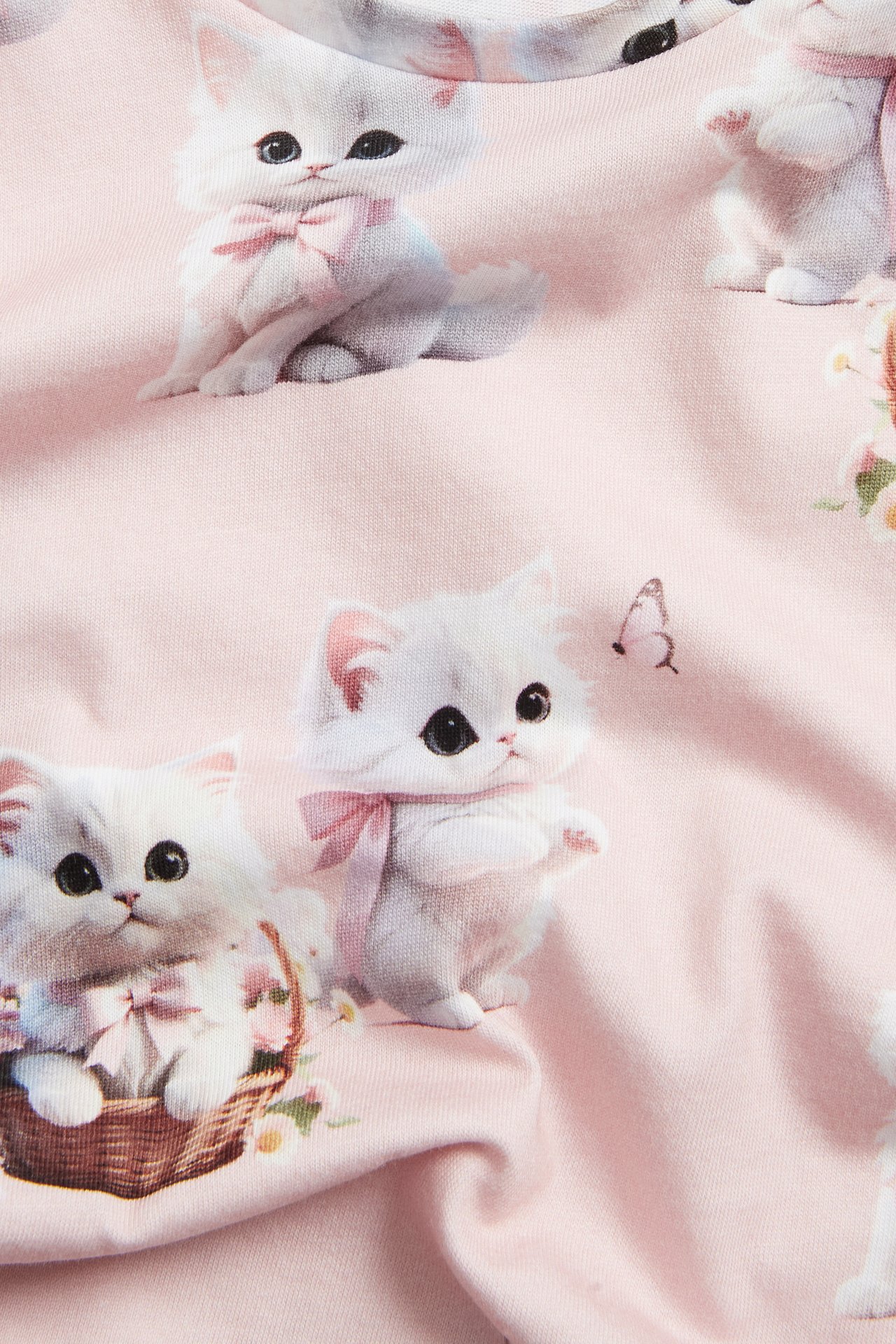 Zbliżenie na jasnoróżowy bawełniany top z krótkim rękawem z nadrukiem białych kotów z różowymi kokardkami.