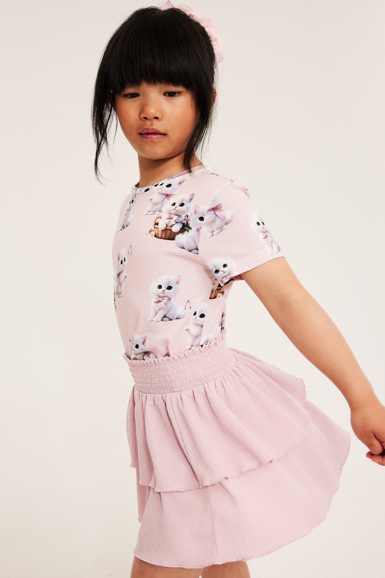 Edestäpäin sivukuva: Lapsi yllään vaaleanpunainen kissakuvioinen puuvilla-T-paita ja röyhelöinen vaaleanpunainen hame.